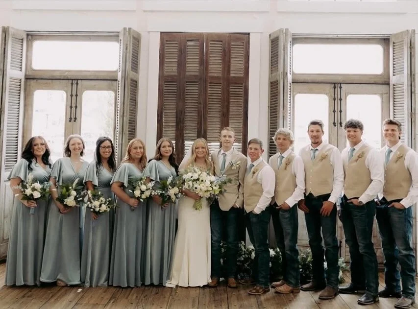 A wedding party standing in front of large windows and wooden shutters, with the bride and groom in the center, surrounded by bridesmaids and groomsmen.