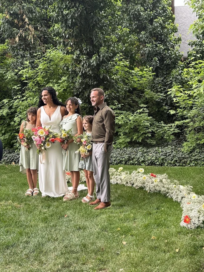 A wedding party standing together outdoors on a grassy area, with lush green trees and bushes in the background. The group includes a woman in a white wedding dress holding a bouquet, two young girls in light green dresses with floral bouquets, and an older man in a brown shirt and light gray pants. The scene is decorated with white and red flowers along the ground.
