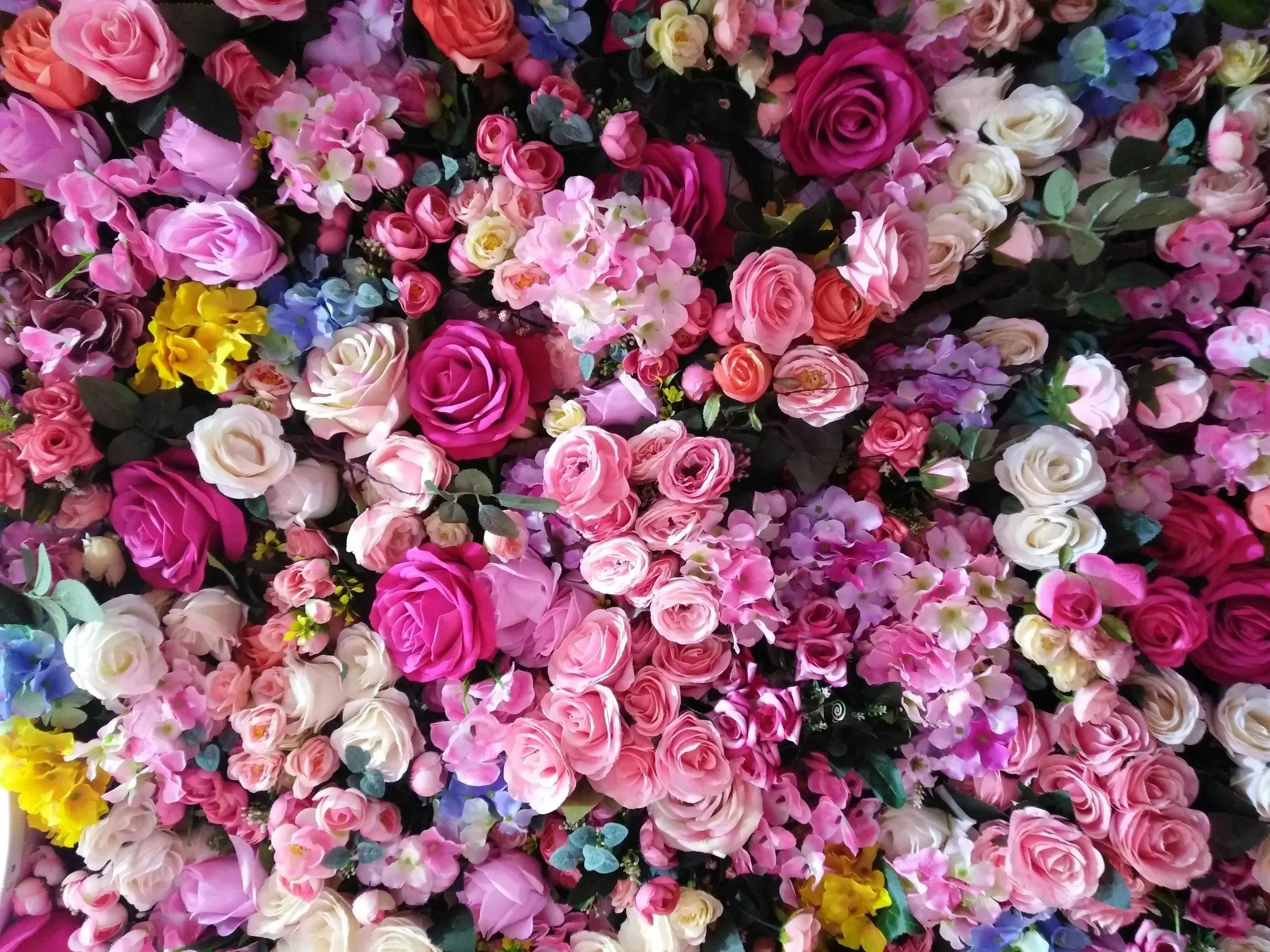 A dense arrangement of colorful artificial flowers, predominantly pink, white, purple, yellow, and blue roses, with green leaves interspersed throughout.