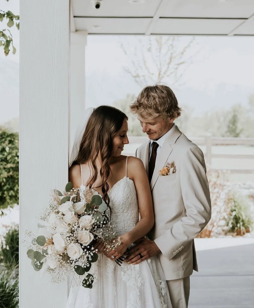 Bride and groom standing close together, smiling, during their wedding, with the bride holding a bouquet of white and blush roses, baby's breath, and eucalyptus.