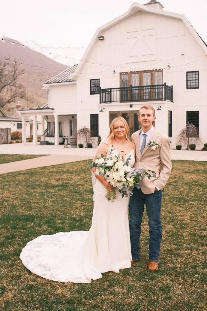 A bride and groom standing on grass in front of a large white barn with a mountain in the background. The bride is holding a bouquet of white flowers and greenery, wearing a white wedding dress with lace sleeves, and the groom is wearing a beige suit jacket, white shirt, gray tie, and dark jeans.