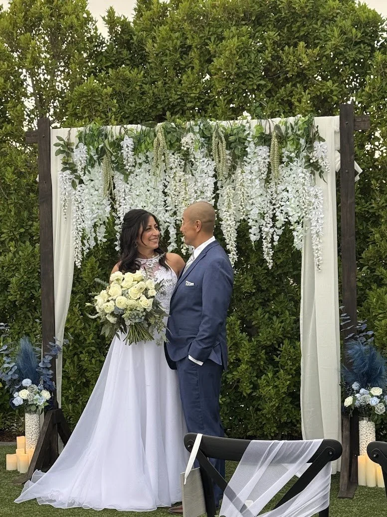 Bride and groom standing close together during their wedding ceremony outdoors, with wedding arch decorated with white flowers and greenery behind them.