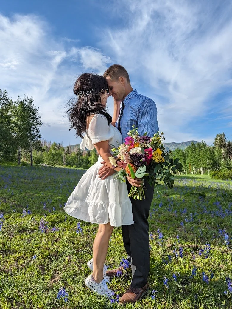 A couple stands close together in a field of purple flowers, with a blue sky and trees in the background. The woman is wearing a white dress and white sneakers. The man is wearing a blue shirt and dark pants, holding a colorful bouquet of flowers. They are leaning their foreheads together, touching in an intimate moment.