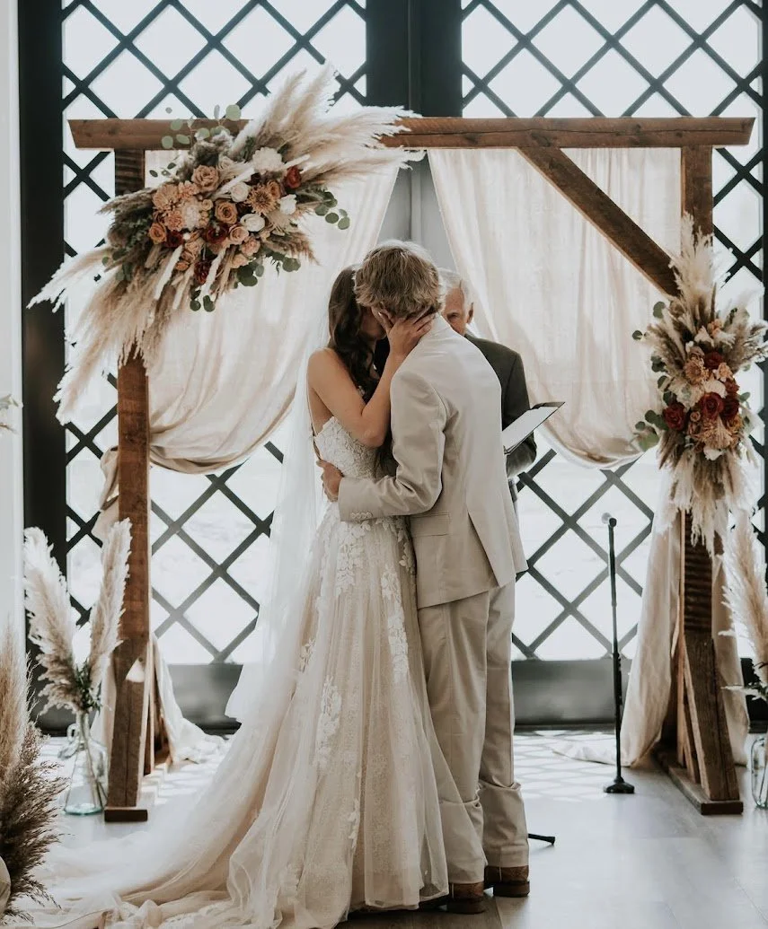 A couple is kissing during a wedding ceremony, standing under a wooden arch decorated with flowers and draped fabric.