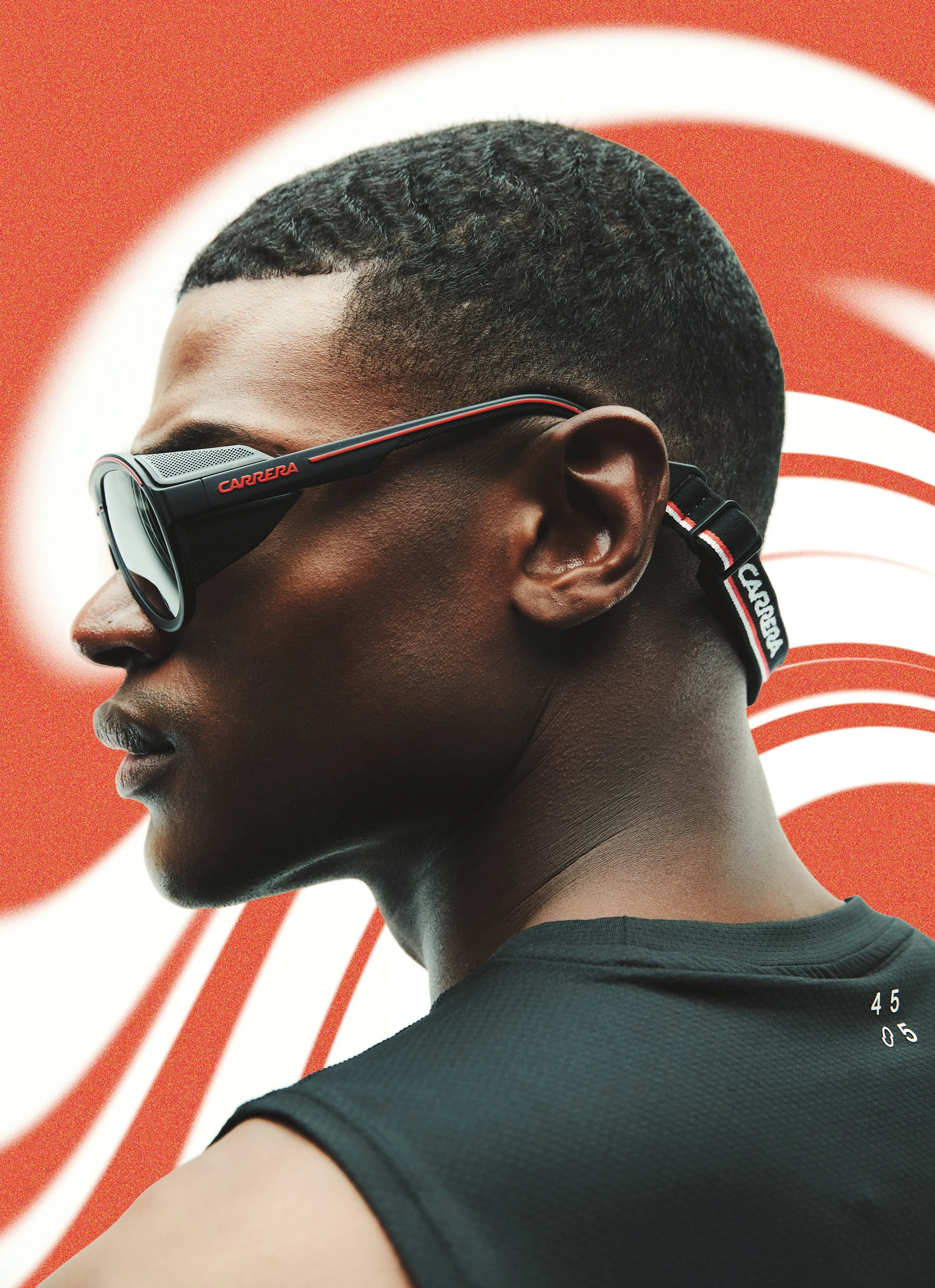 Close-up of a man's profile with short, textured black hair, wearing sporty sunglasses with 'CARRERA' branding, and a black athletic shirt with a round neckline. The background features a red circle with white lines radiating outward, creating a dyna