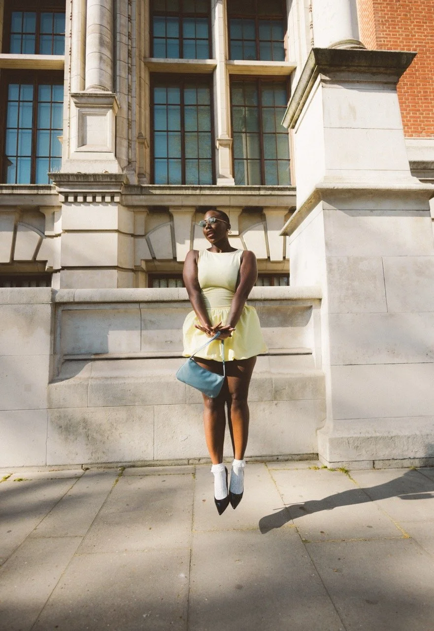 A woman in a yellow dress appears to be floating above the ground, with her shadow visible on the sidewalk, against a historic building with large windows.