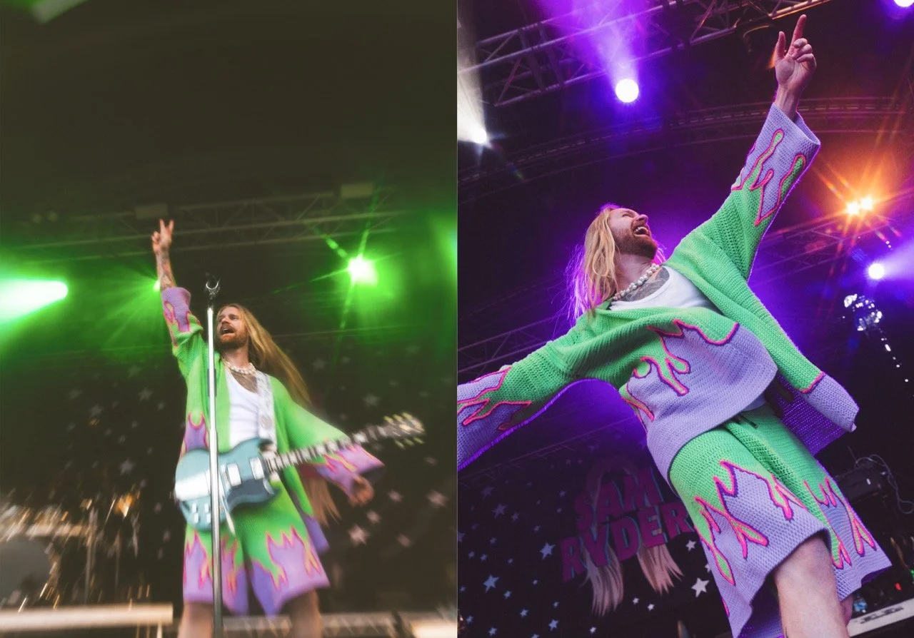 Performer on stage wearing a colorful green and pink outfit, playing guitar and singing with lights and stage equipment in the background.