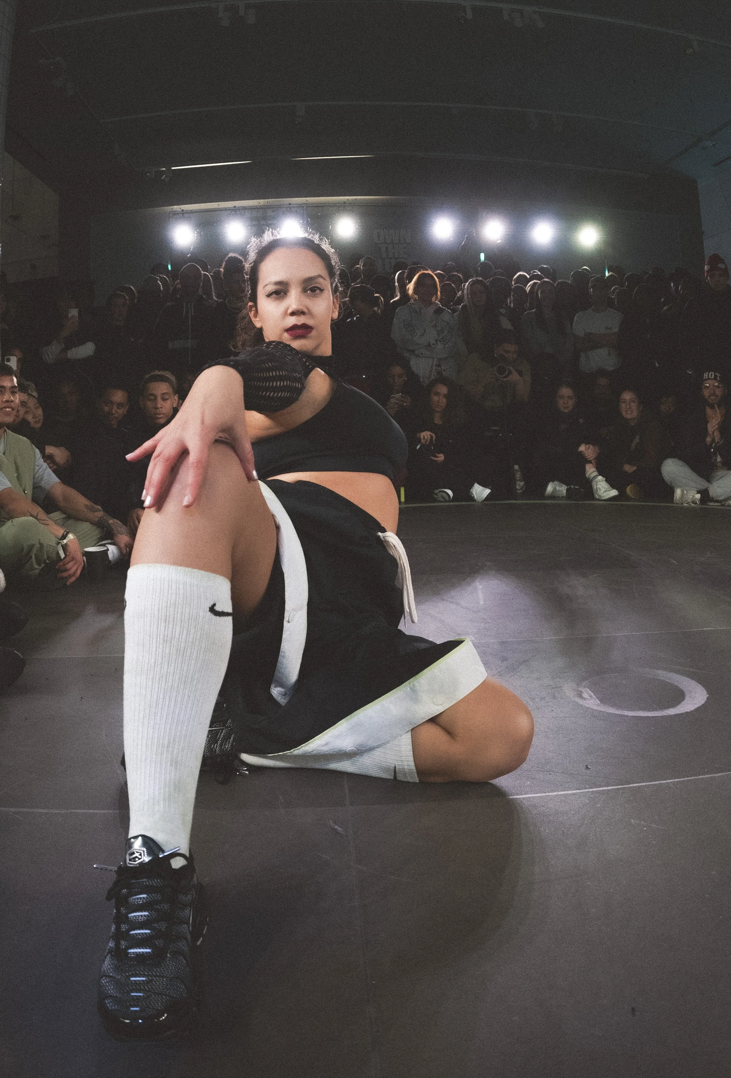 A woman kneeling on the floor in a dark room with a large audience and bright stage lights in the background. She is wearing a black sports top, Nike shorts, knee-high white socks, and black sneakers. She has dark hair tied back and dark lipstick.