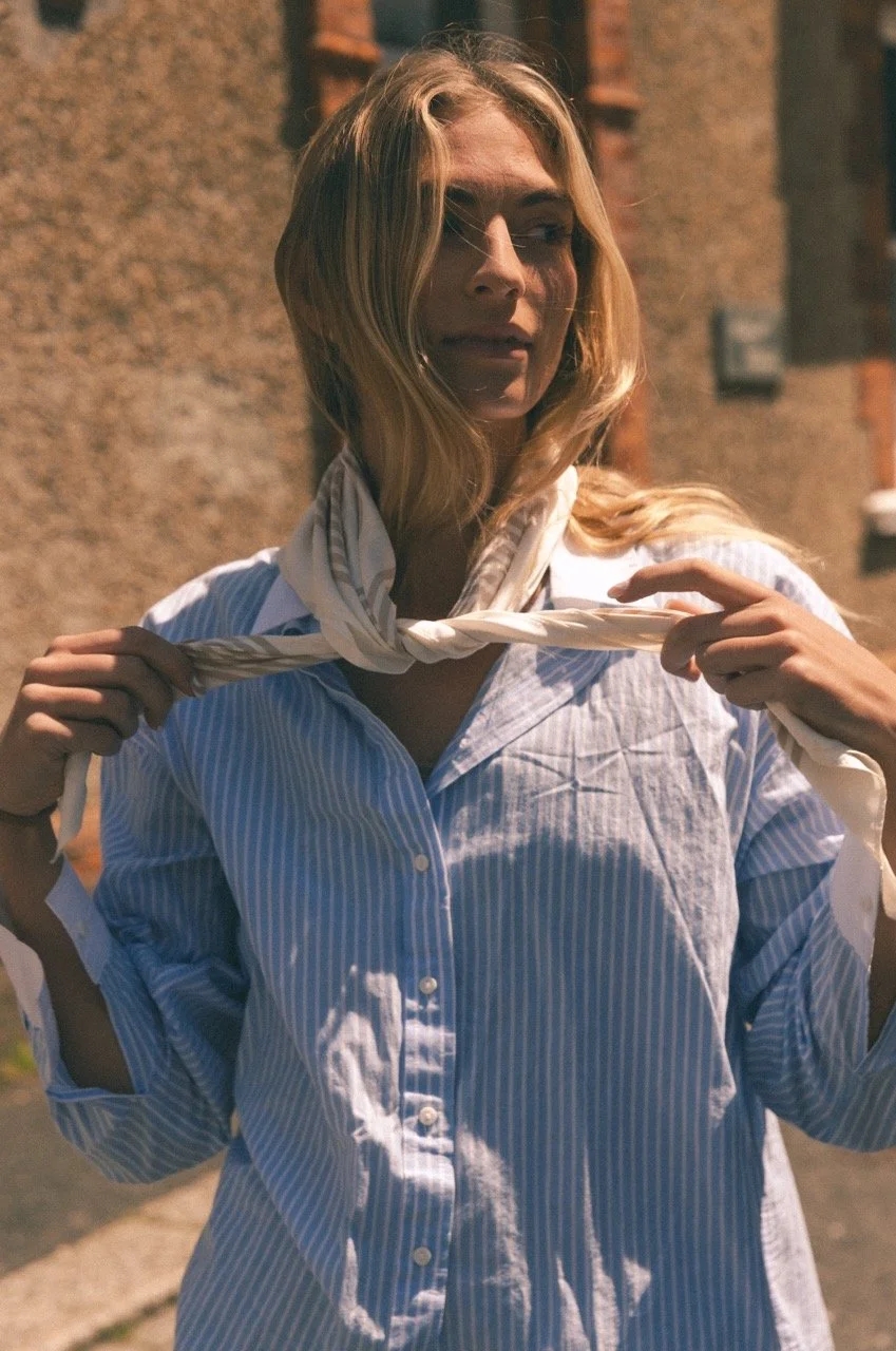 A woman with blonde hair, wearing a striped shirt, is outdoors at sunset, holding a white scarf around her neck, with a brick wall behind her.