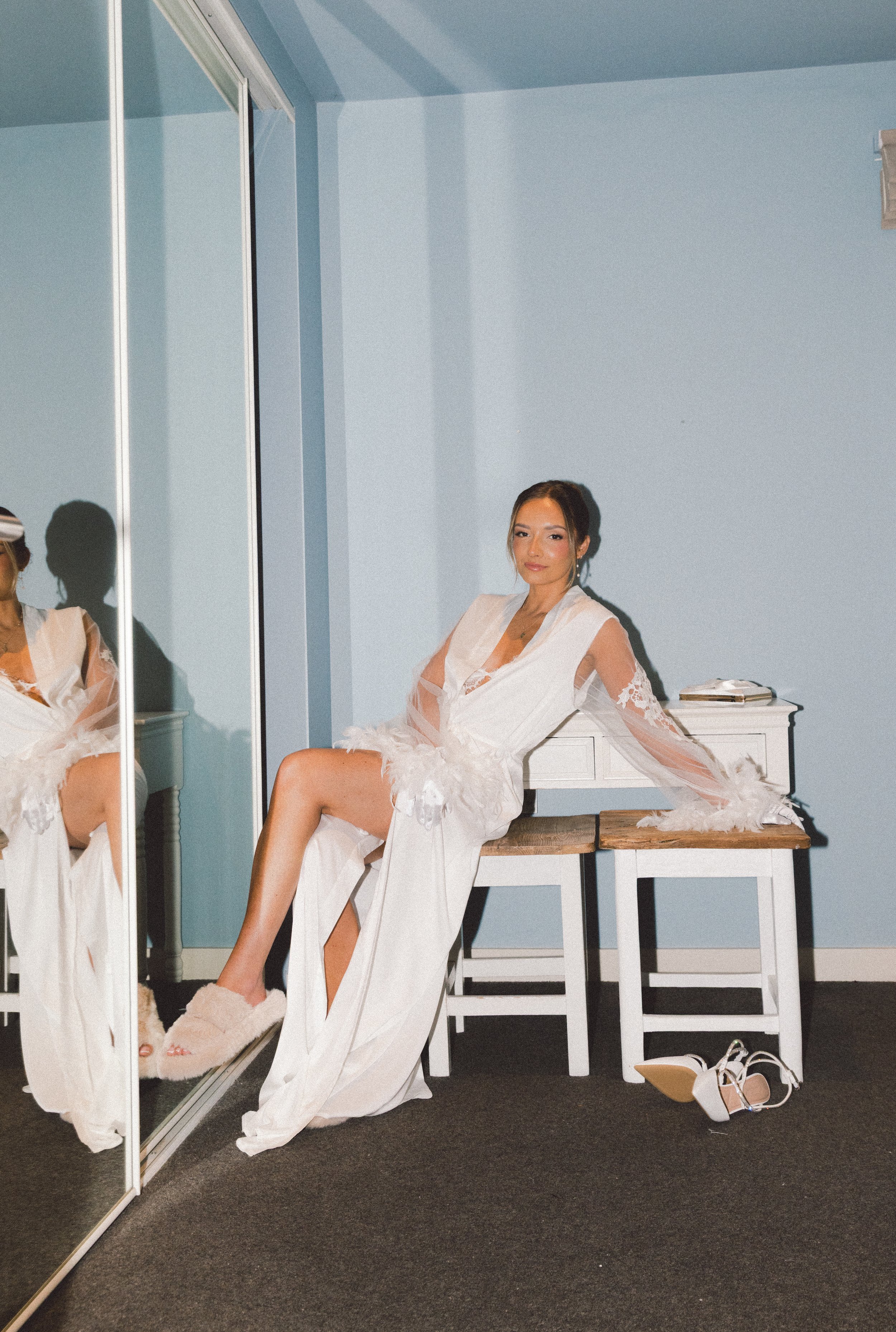 A woman in a white dress with lace details sitting on a chair in a room with light blue walls, a large mirror, and shoes on the floor nearby.