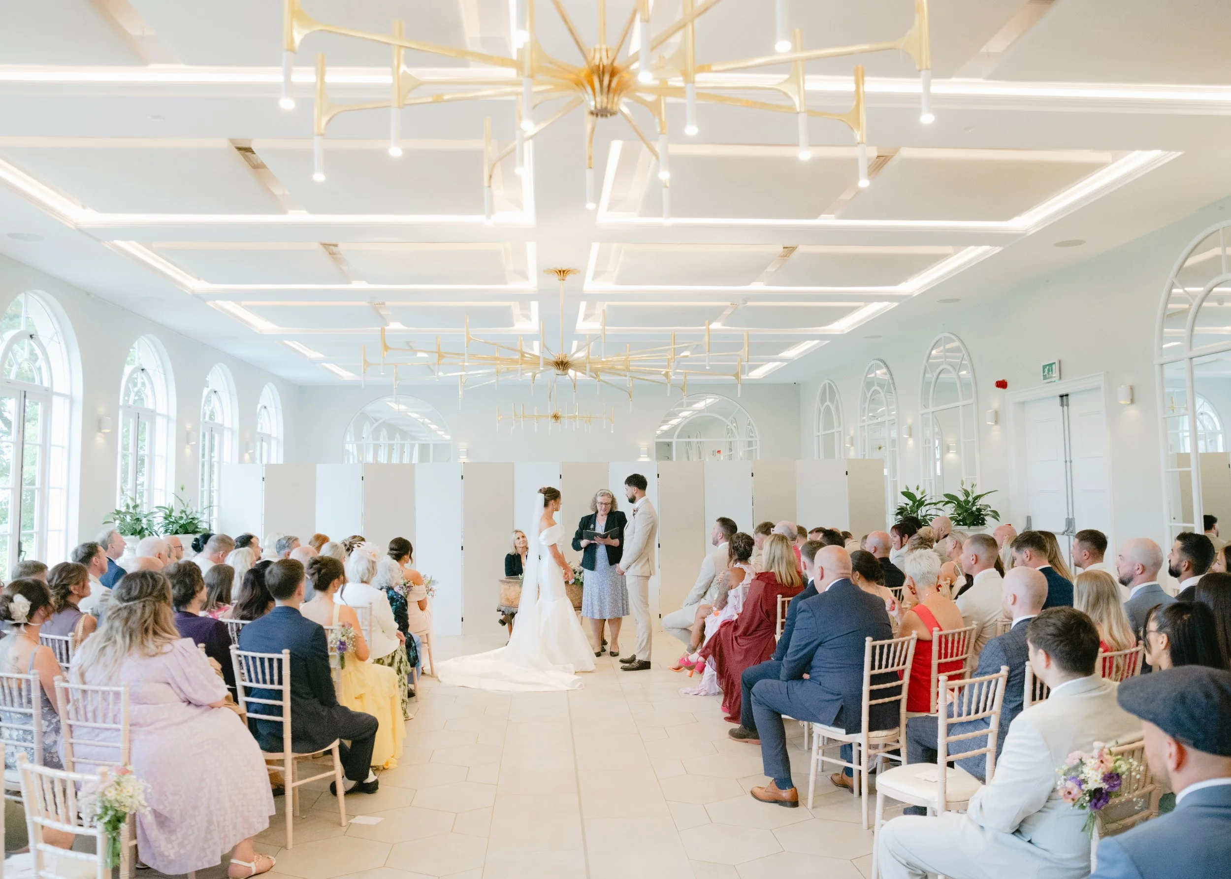 A wedding ceremony inside a bright, modern hall with large arched windows and decorative chandeliers. The bride and groom are standing in front of an officiant, holding hands while guests watch and seated on either side.