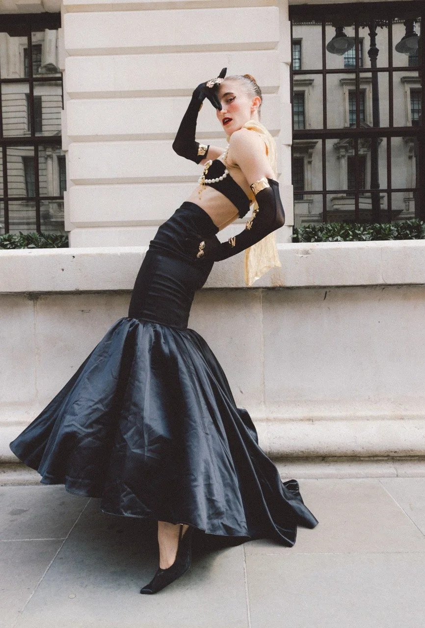 A woman in a fashionable black and gold outfit, wearing long black gloves and high heels, posing against a city building background.
