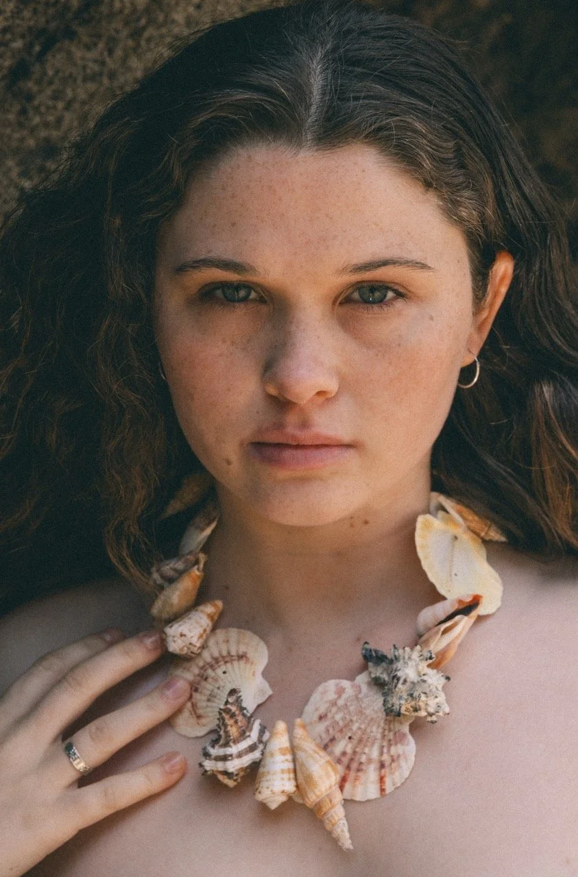 A young woman with light skin and dark hair lying down outdoors wearing a seashell necklace and a ring on her finger.
