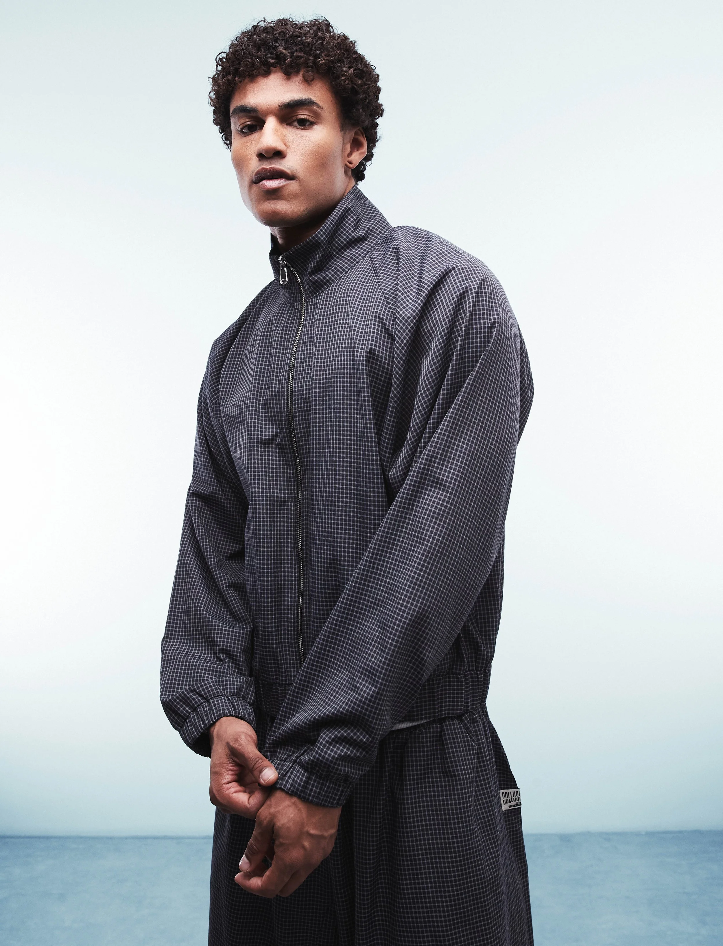 A young man with curly hair wearing a dark checkered zip-up jacket and matching pants standing outdoors against a background of cloudy sky and ocean.
