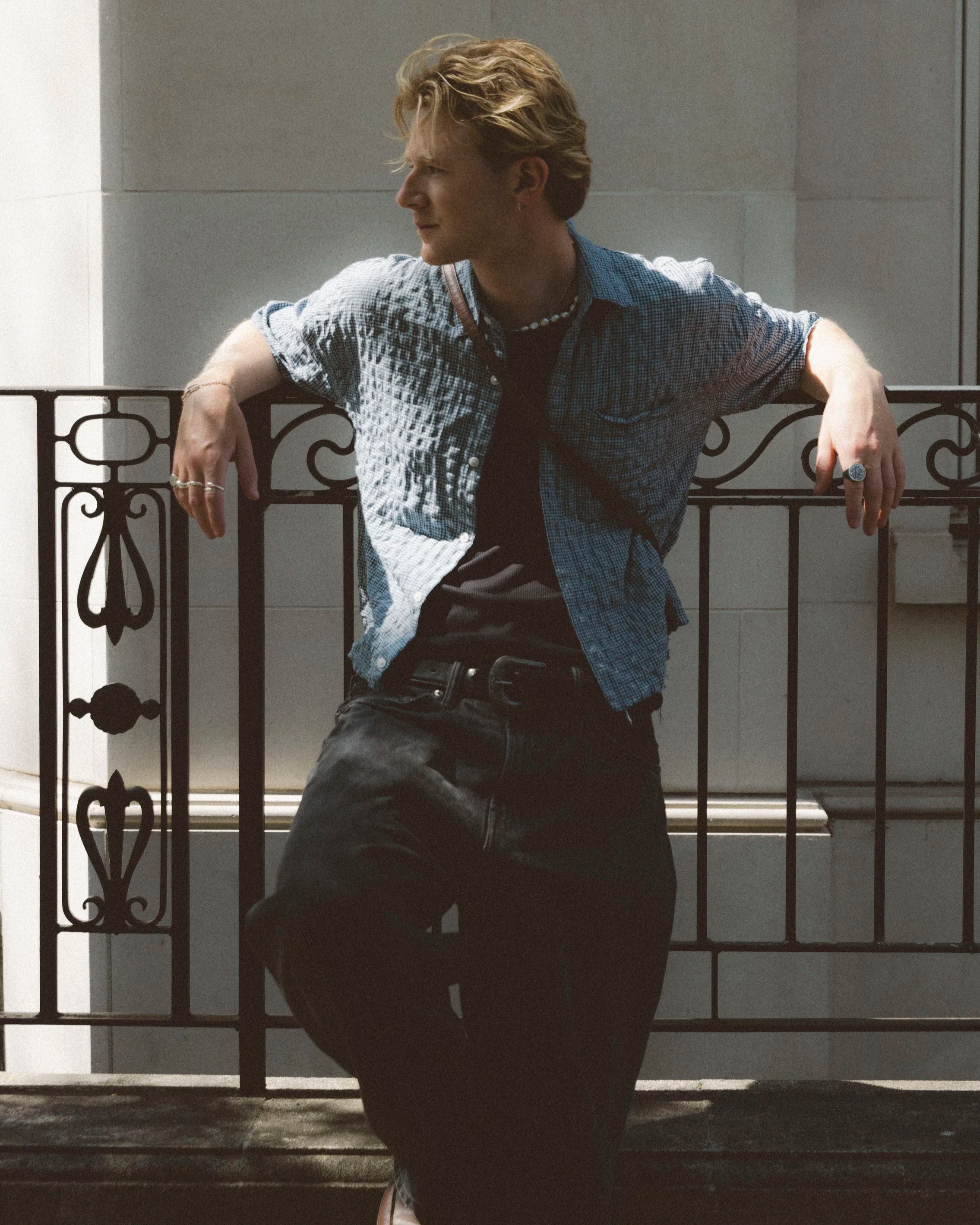 A young man with blond hair, wearing a checkered shirt and black pants, leaning on a black decorative metal railing outside a building with a light-colored wall.