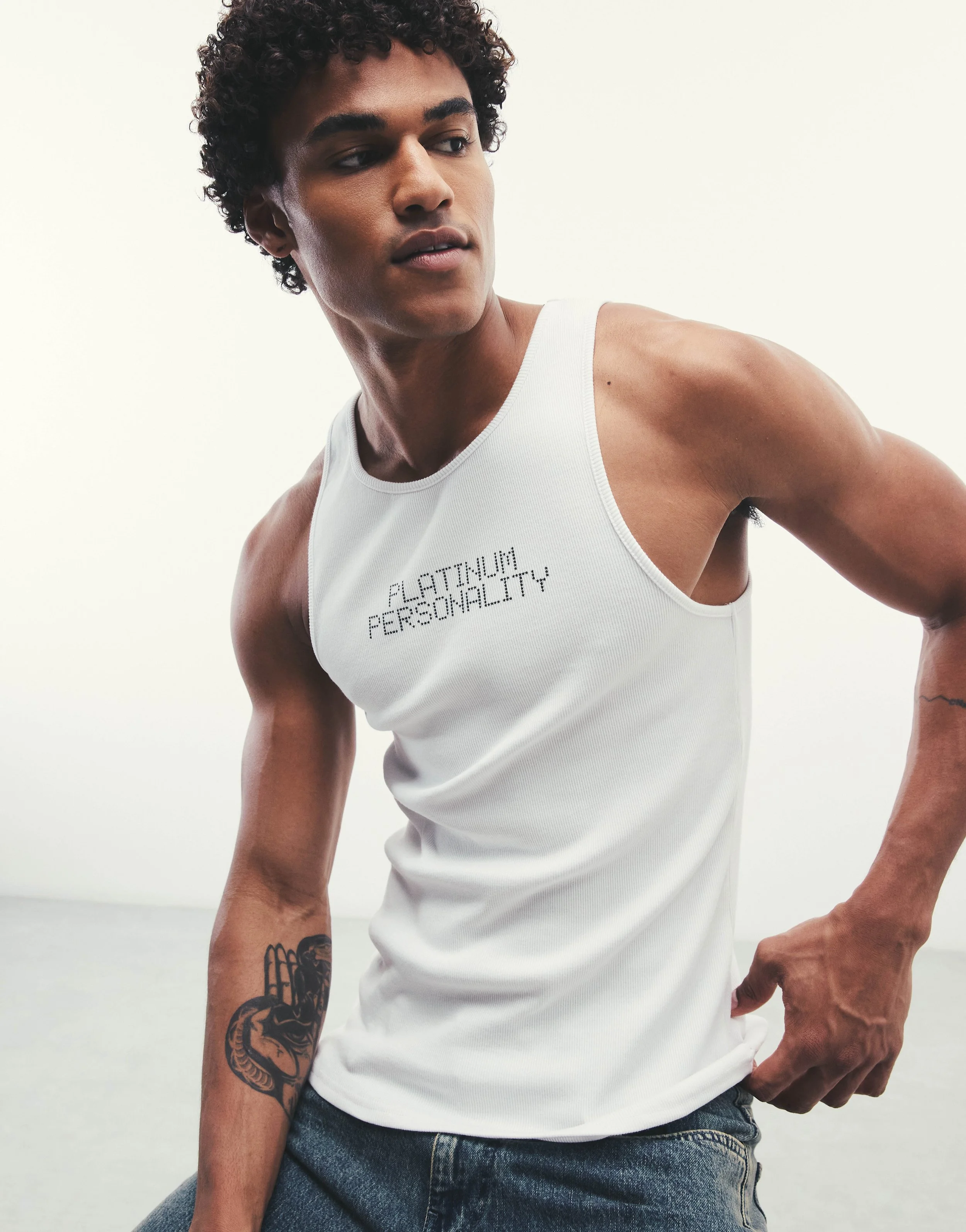 A young man with curly hair wearing a white tank top and jeans, showing a tattoo of a dragon on his left forearm, standing against a plain white background.