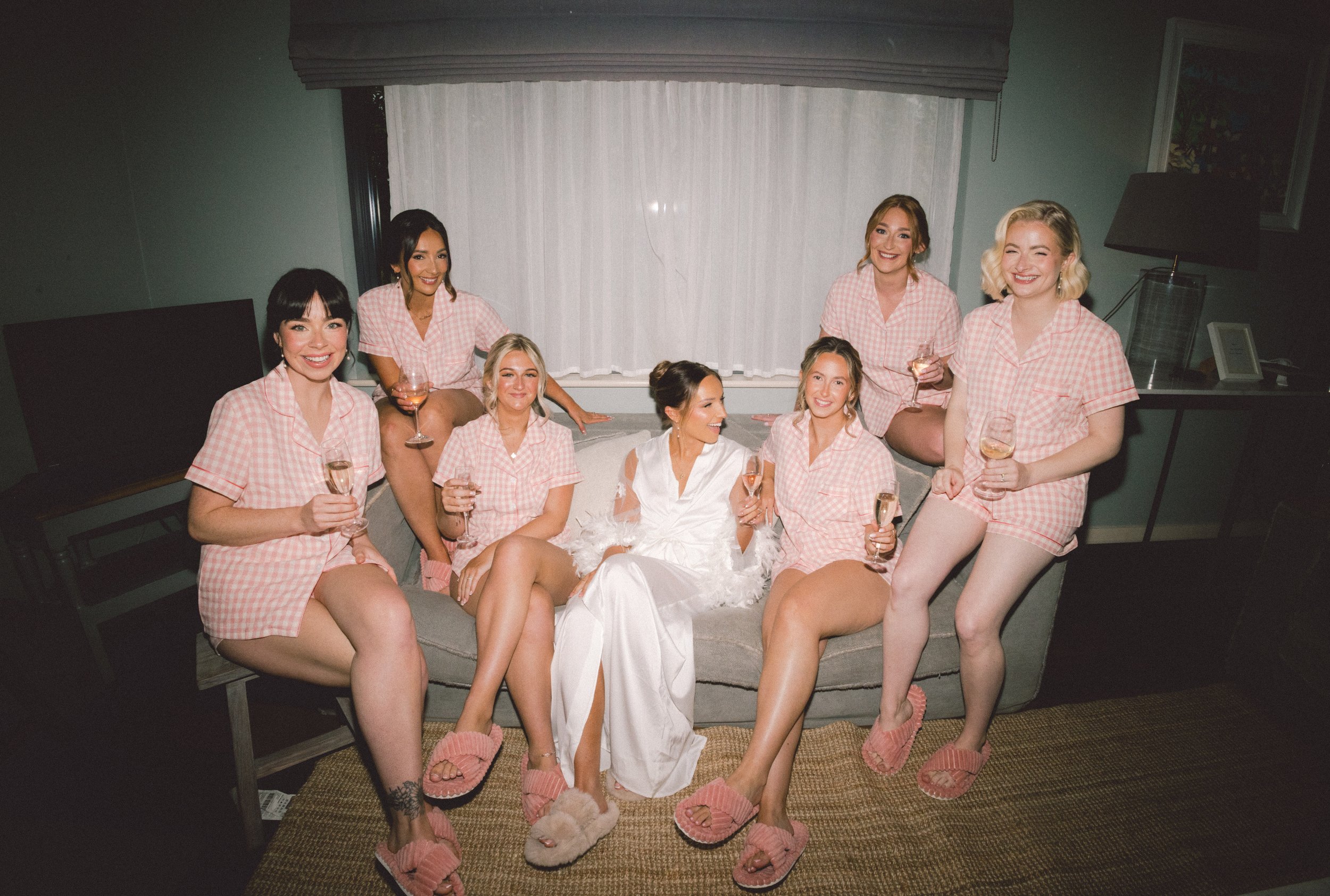 Nine women gathered in a room, dressed in matching pink and white checkered pajamas, holding glasses of wine, and smiling for the photo. They are seated on and around a bed with a large window with white curtains behind them, and some have slippers on.