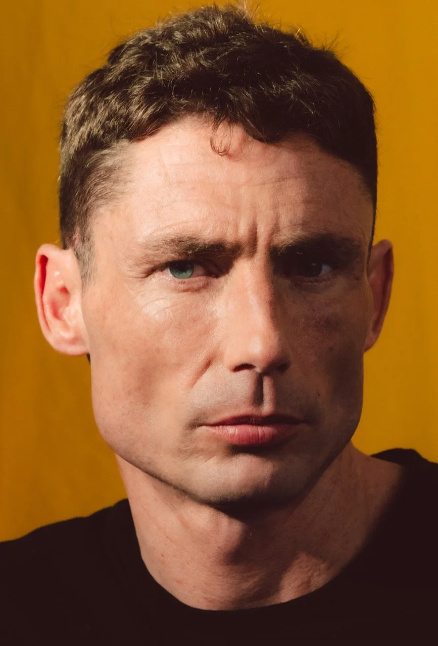 Close-up of a man with short dark hair, blue eyes, and a serious expression against a yellow background.