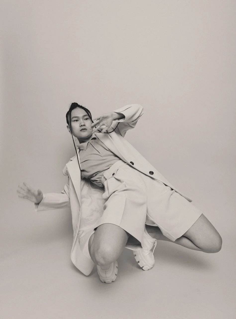 Black and white photo of a young woman in casual fashion striking a pose, with one hand near her face and the other extended outward, against a plain light background.