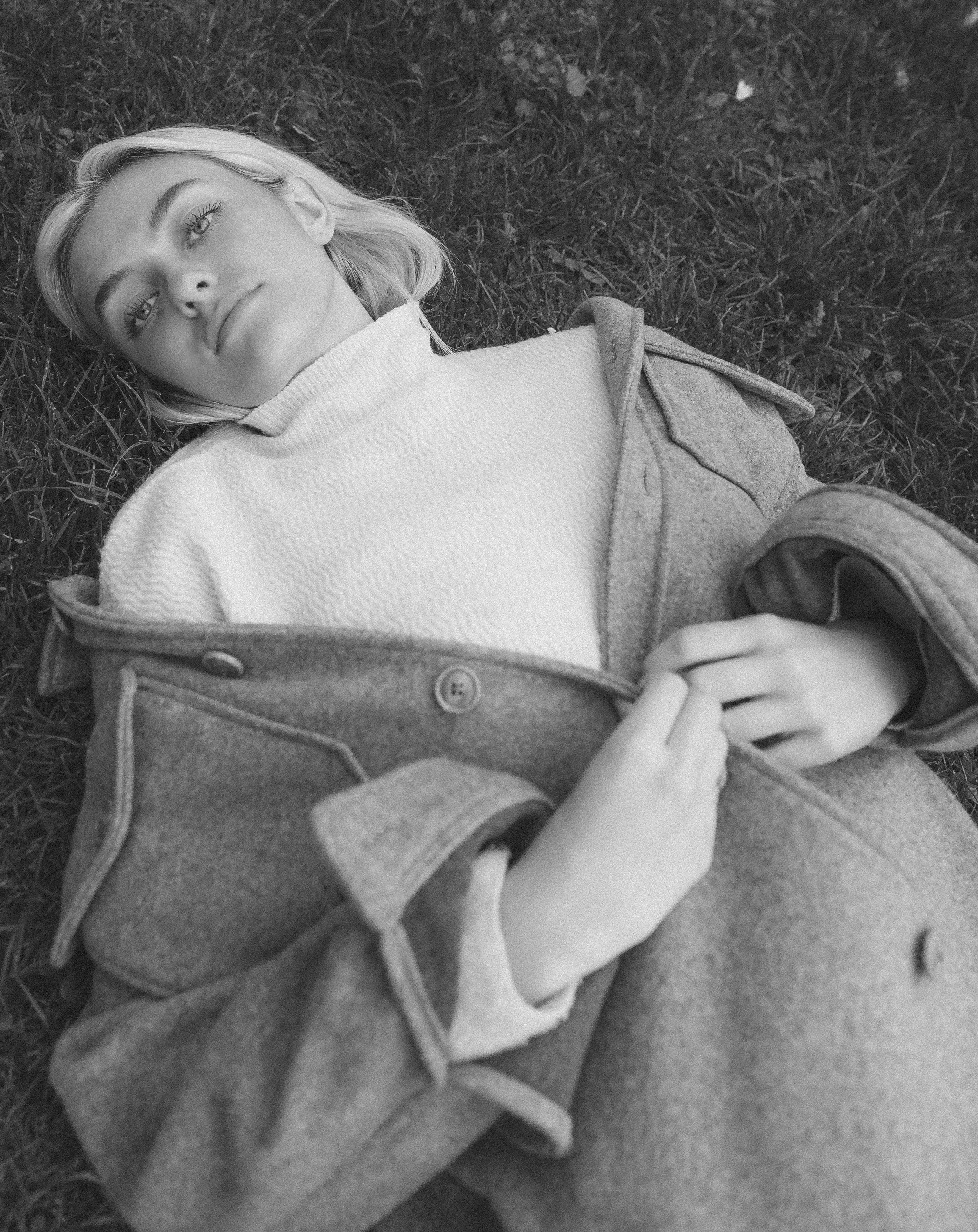 A woman lying on her back on grass, looking up, wearing a light coat and gloves.