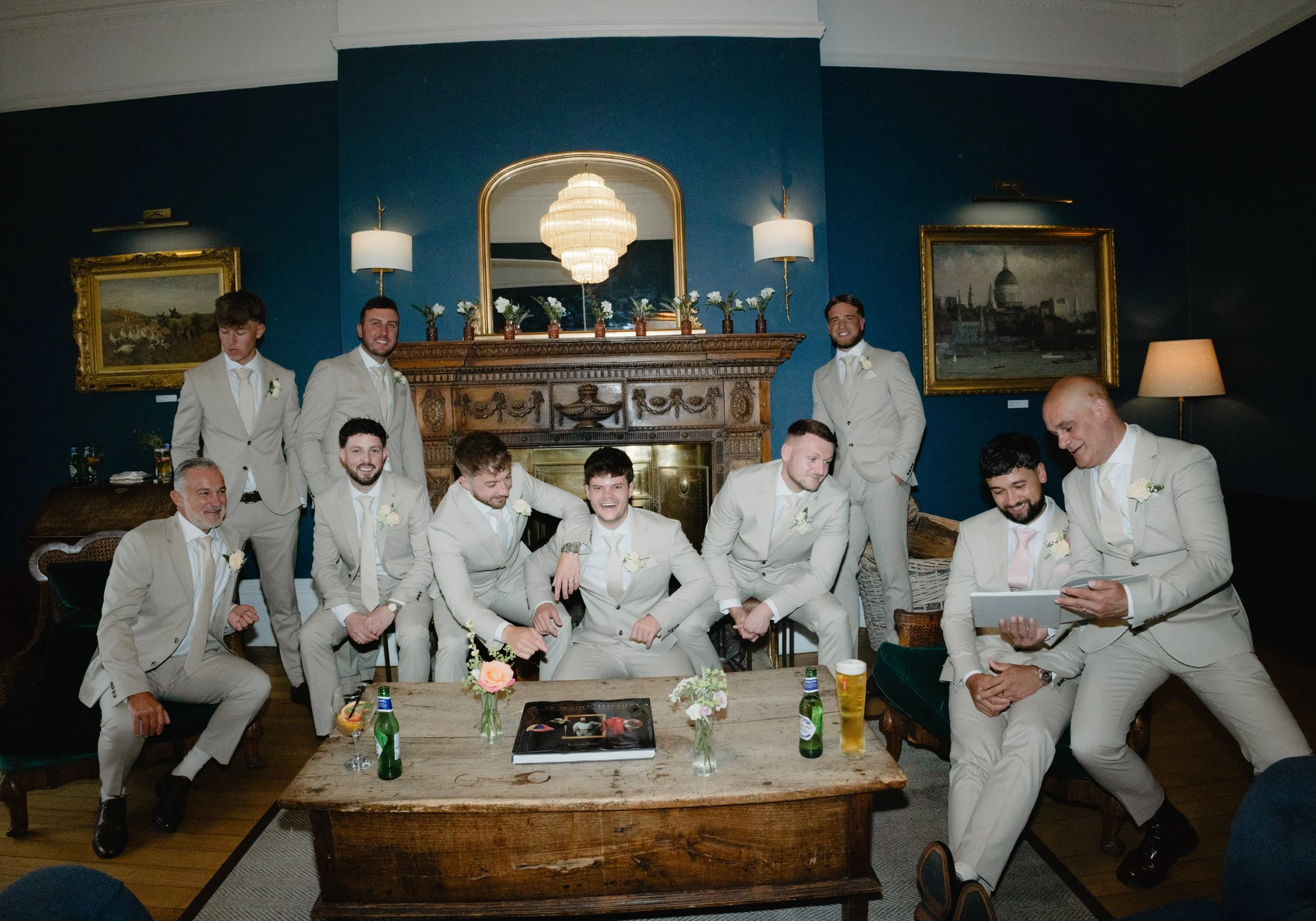 Group of men in beige suits celebrating indoors, sitting and standing around a wooden table with beers and flowers, with a fireplace, paintings, and chandelier in the background.