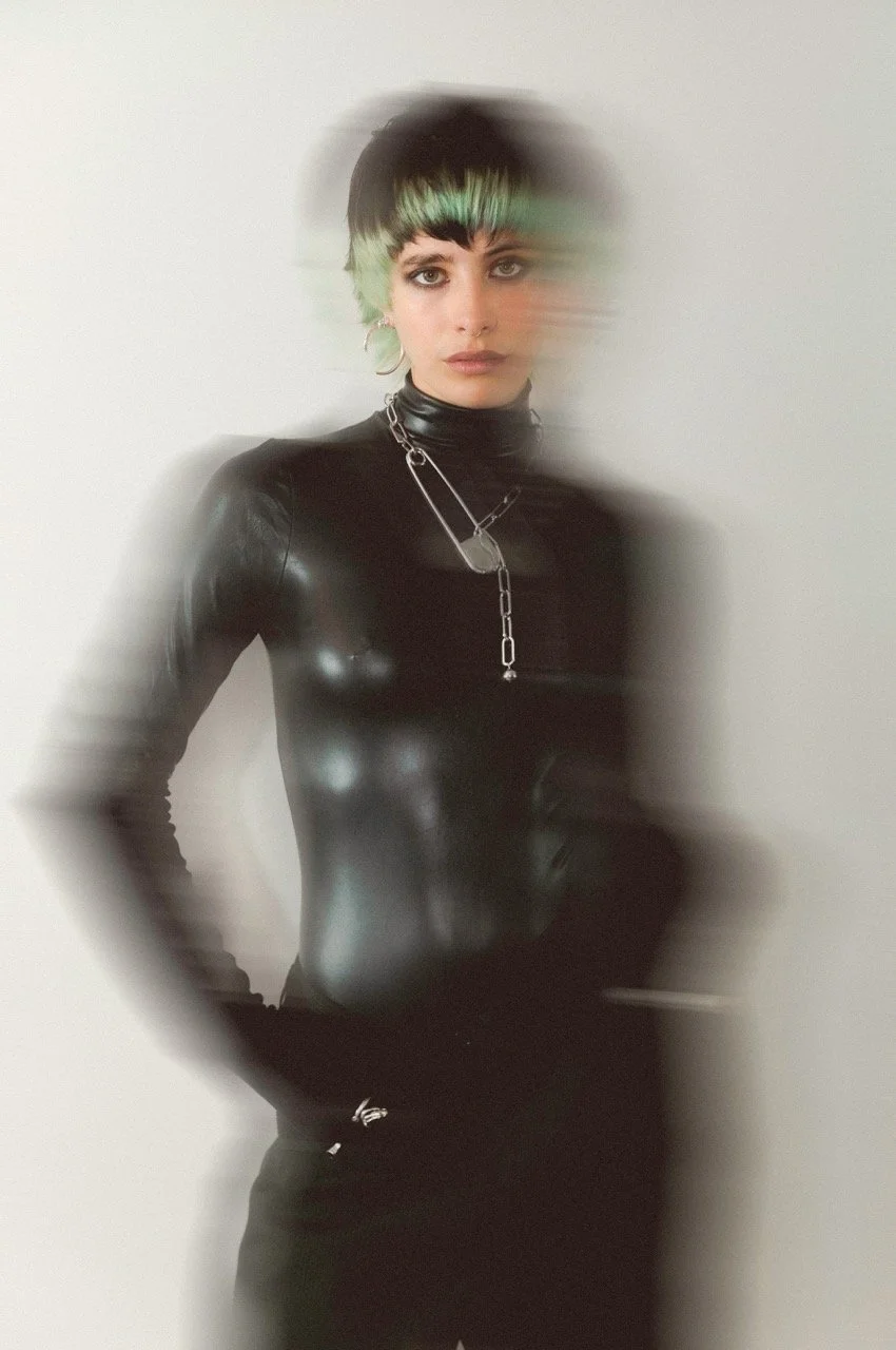 A woman with short black hair with green highlights, wearing a black leather turtleneck and black pants, with layered silver necklaces, standing against a plain light-colored background with a motion blur effect.
