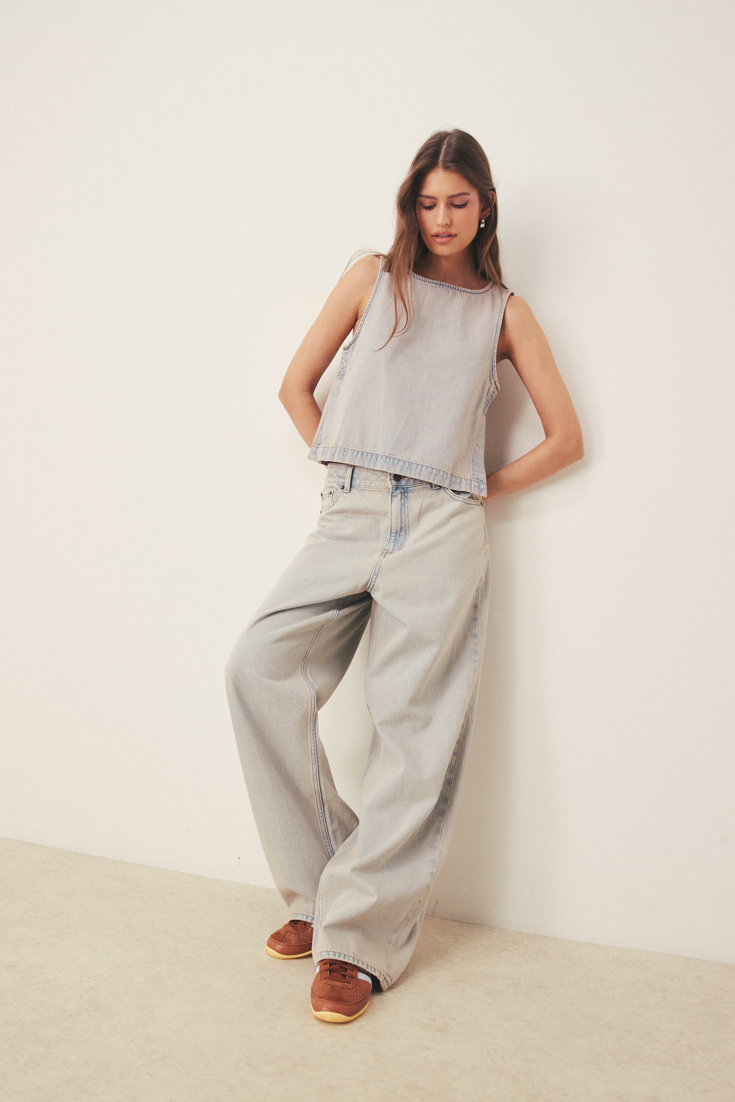 A young woman with long brown hair and light skin is leaning against a plain white wall, wearing a sleeveless denim top and baggy light-colored jeans, with brown sneakers, and looking down.