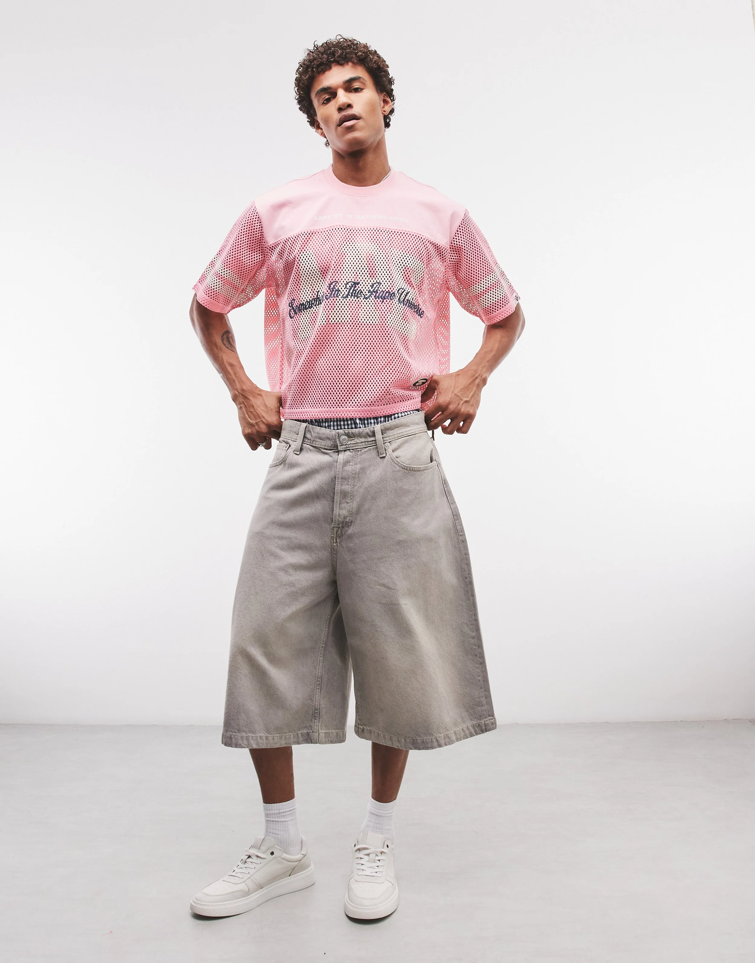 Person wearing a pink mesh t-shirt with text and graphics, gray wide-leg shorts, white sneakers, and white socks, standing against a plain white background.