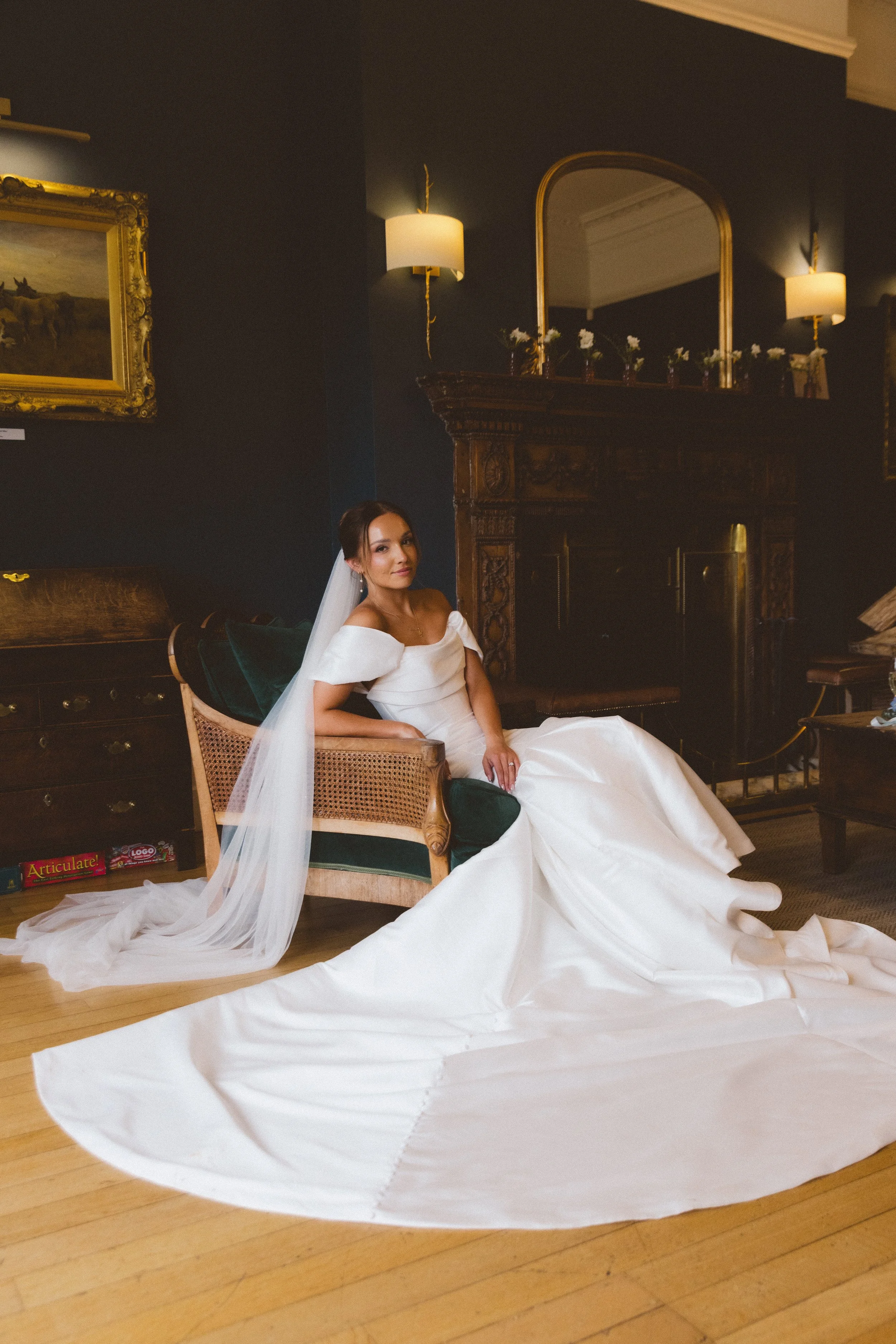 A woman in a wedding dress with a veil sitting on a vintage wooden chair in a dimly lit room with dark walls, a painting, a large ornate mirror, and a wooden fireplace mantle with small flower arrangements.