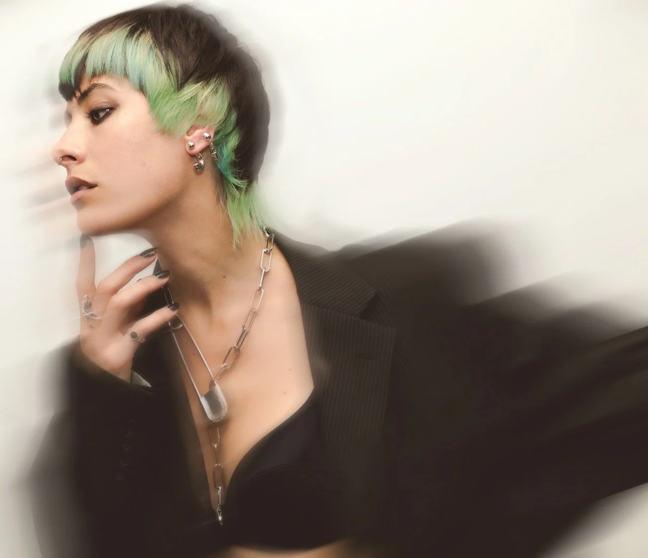 A woman with short hair dyed green and black, wearing multiple earrings, a chain necklace with a large dog tag, and a black blazer, posing with her hand near her face against a plain light background.