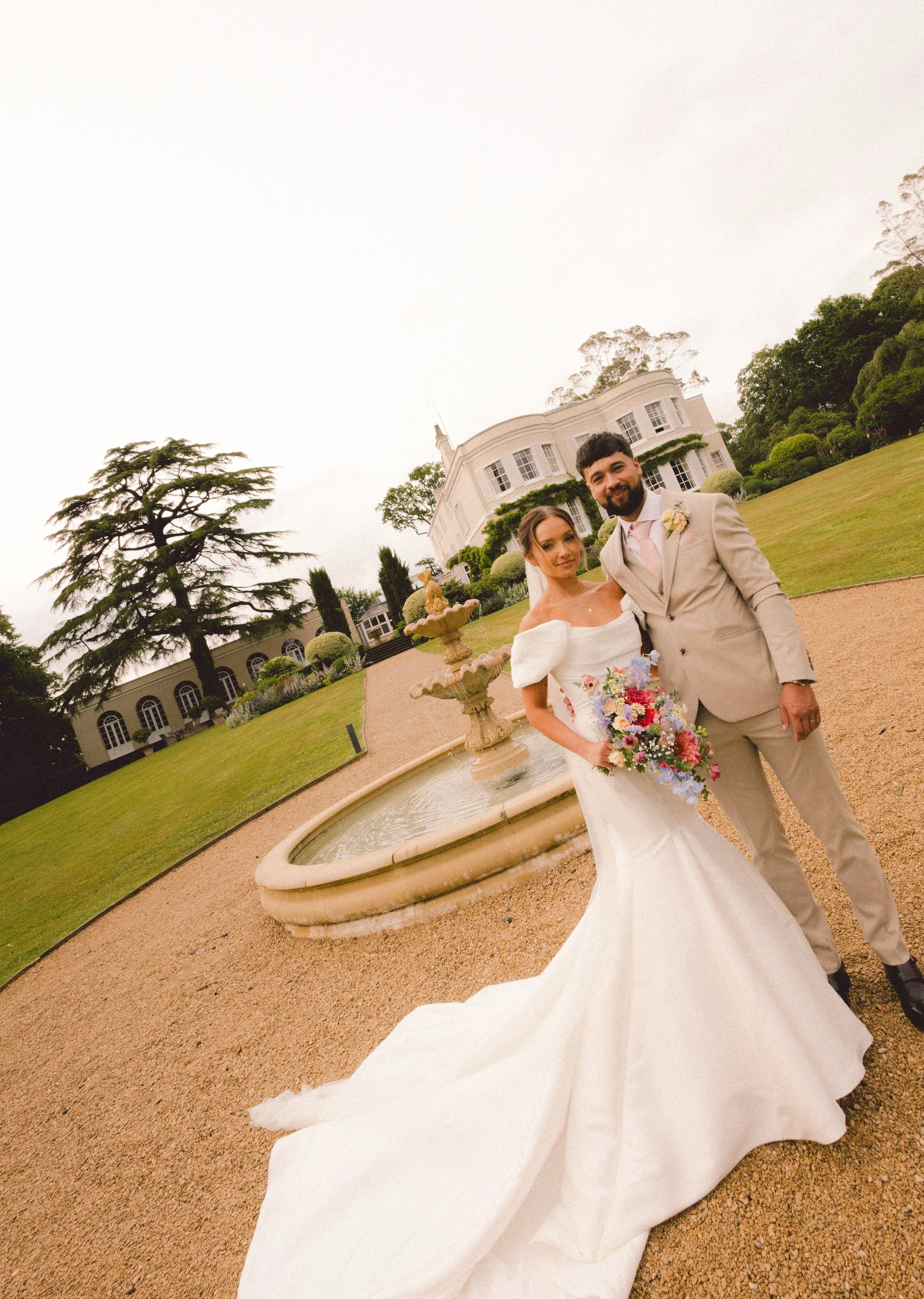 A bride and groom in wedding attire standing outside near a fountain and large white house with greenery, trees, and a cloudy sky in the background.
