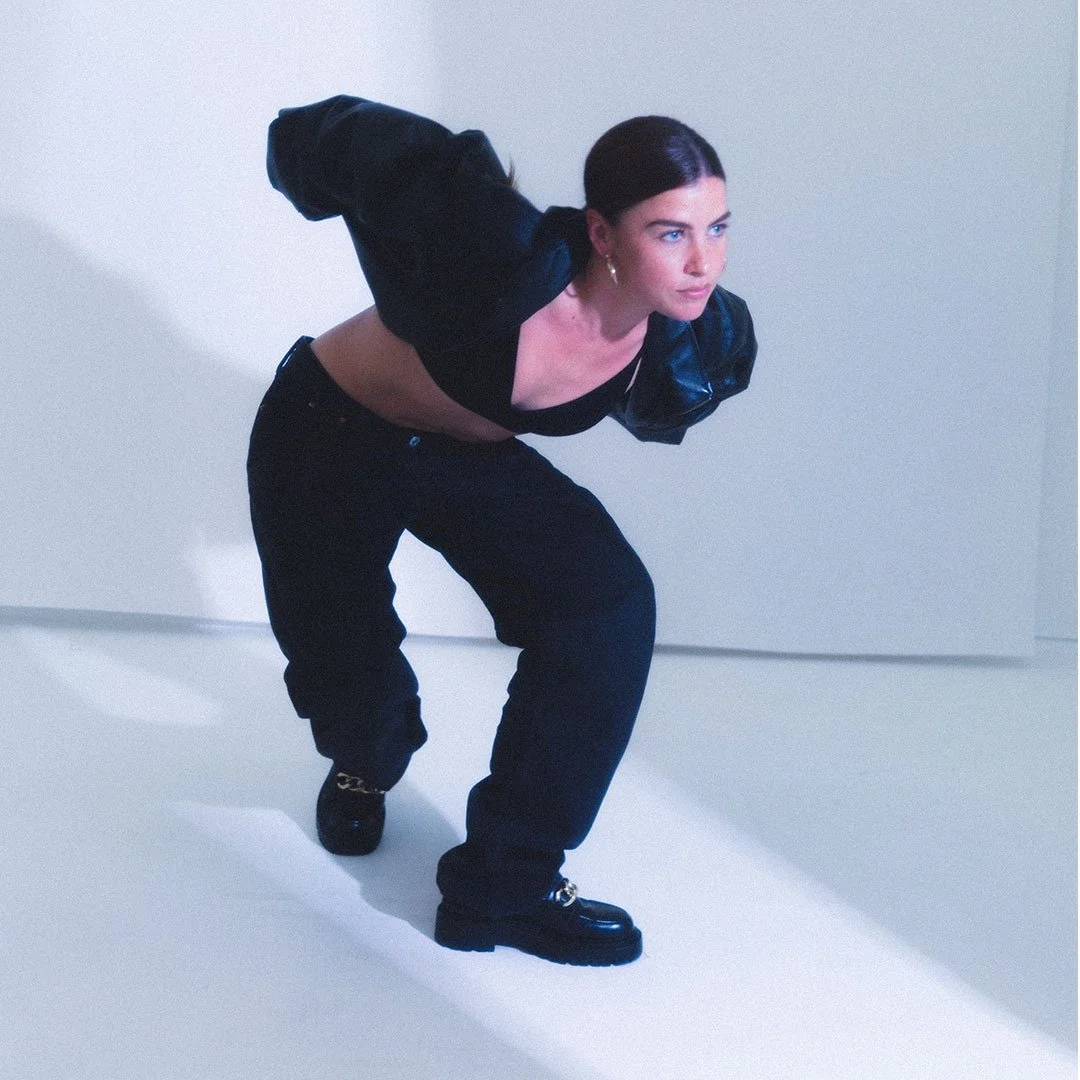 A woman with dark hair wearing a black crop top, oversized black pants, a black jacket with puffed sleeves, and black shoes, crouching in a pose against a plain light background.