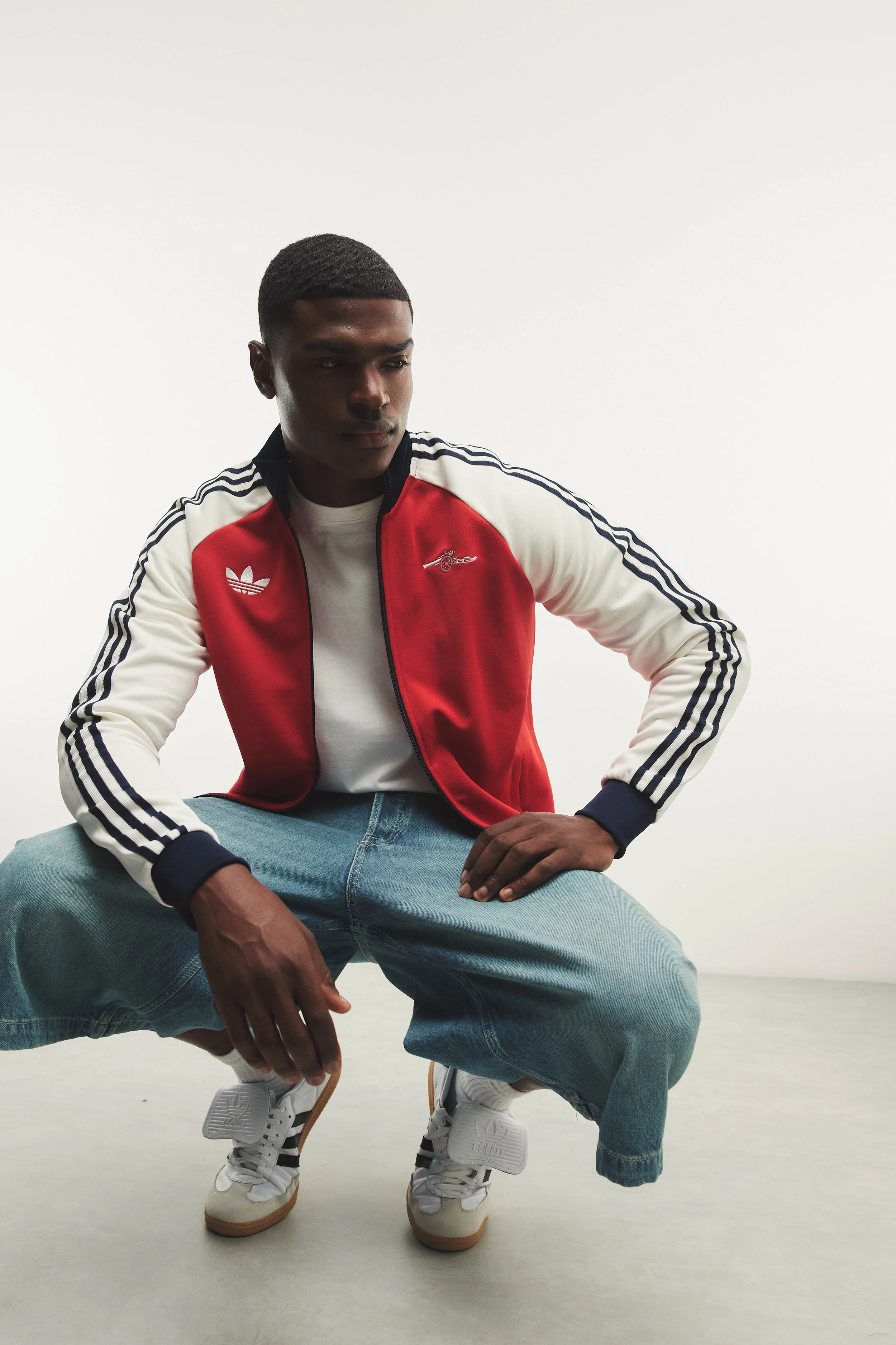A young man with dark skin and short black hair styled in waves, wearing a red and white Adidas track jacket with black stripes, a white t-shirt, loose blue jeans, and white sneakers with black accents. He is squatting down with his hands resting on 