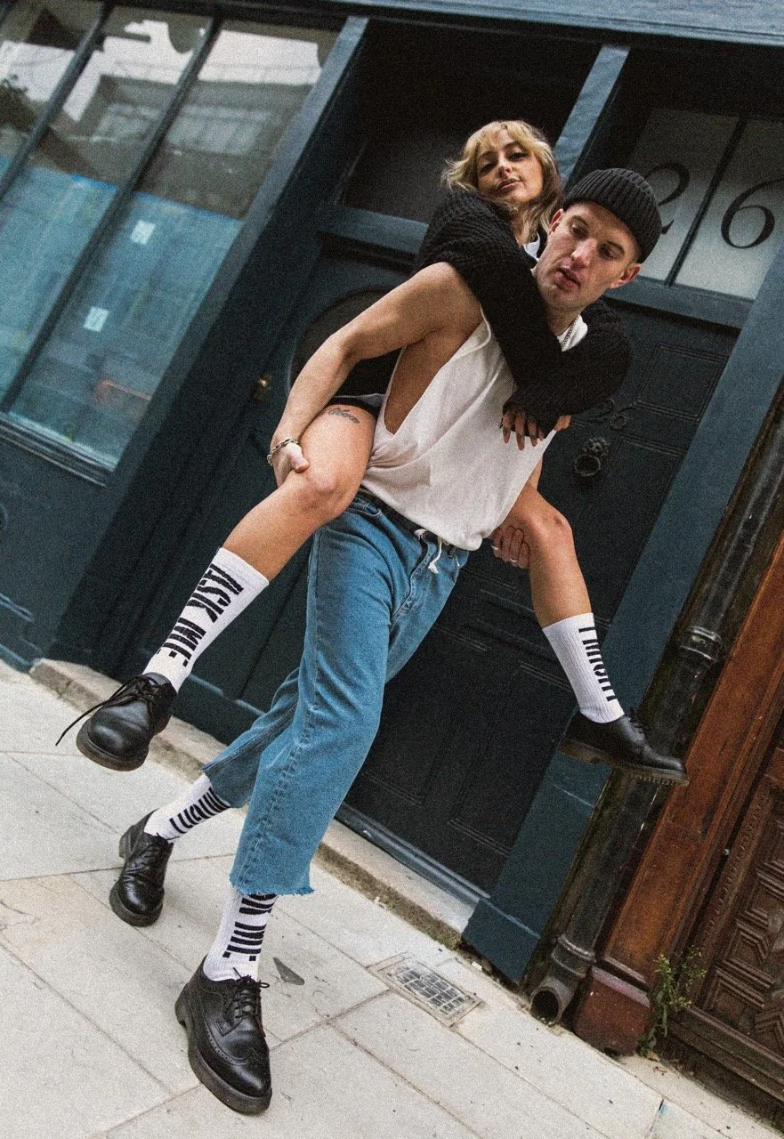 A young man carrying a woman on his back outside a building with dark blue door and window frames. The woman is wearing black socks and black shoes, while the man is dressed in casual jeans, a sleeveless shirt, and a beanie. They are posing playfully