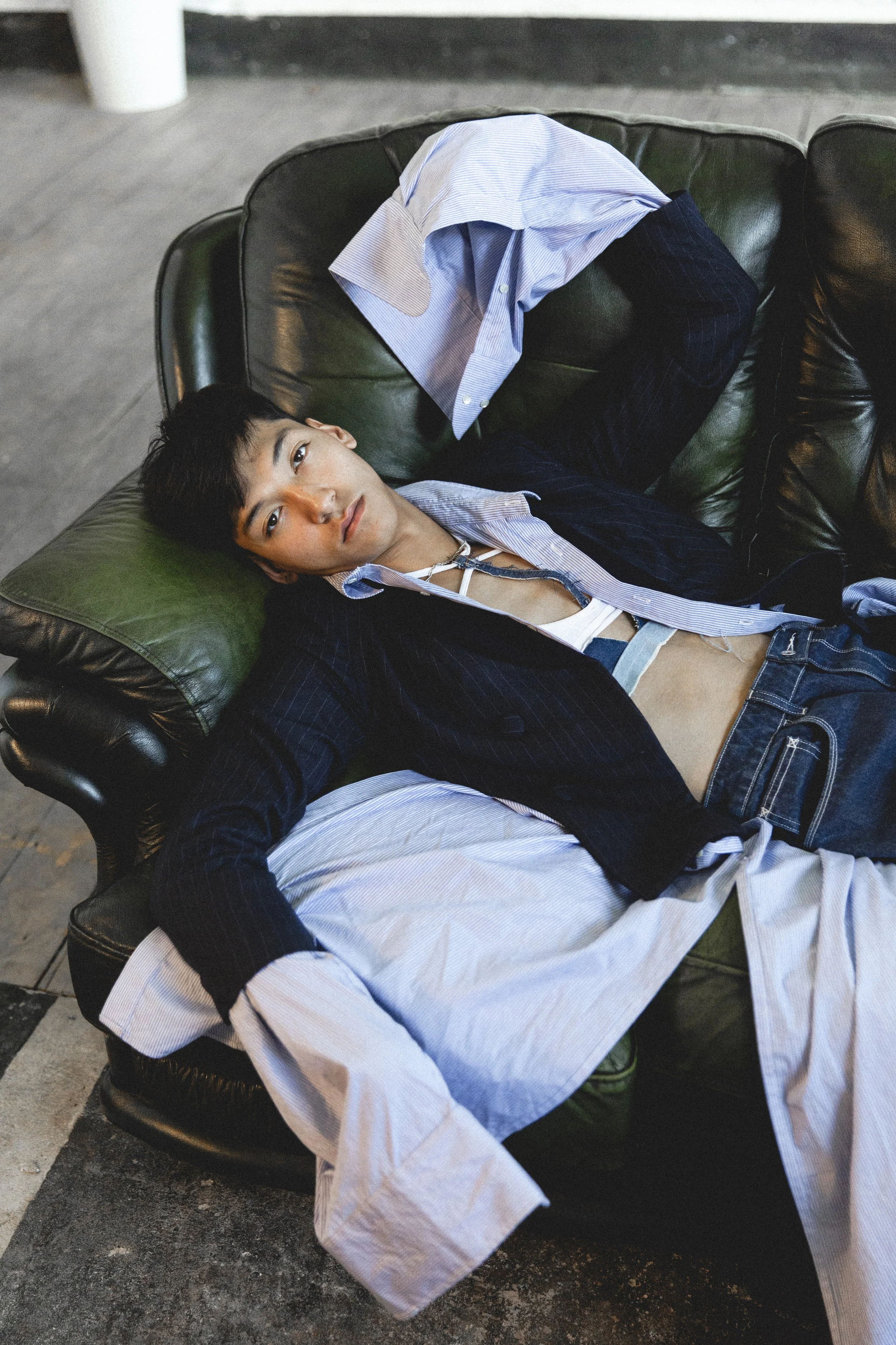A young man with dark hair and light skin lying on a green leather couch, wearing a partially unbuttoned white shirt under a dark blazer, with an open collar and a white zipper top underneath. A white shirt is draped over the cushion and his arm. He 