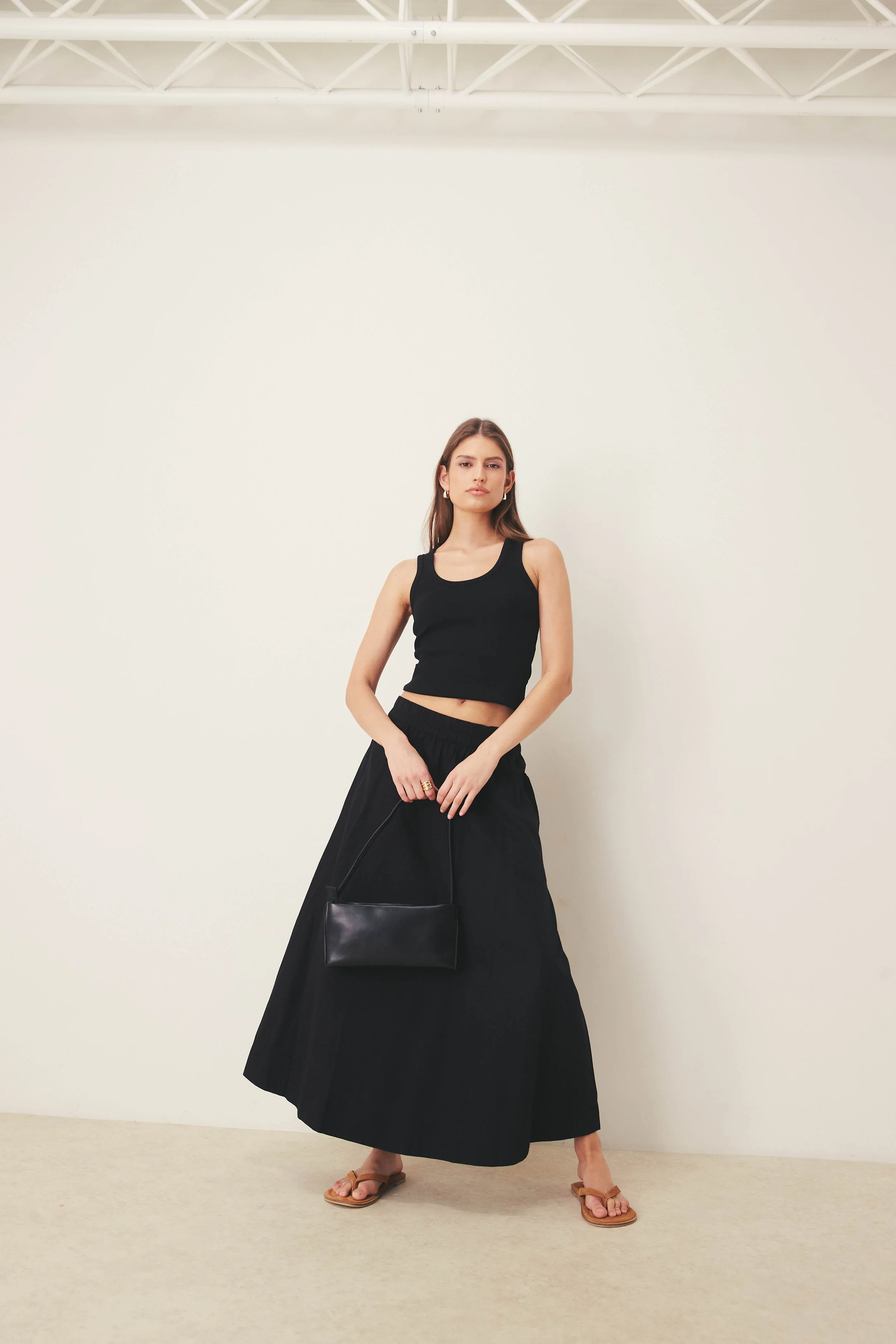 A young woman with long brown hair standing in front of a plain white background, dressed in a black sleeveless crop top and a long black skirt, holding a black purse and wearing tan sandals.