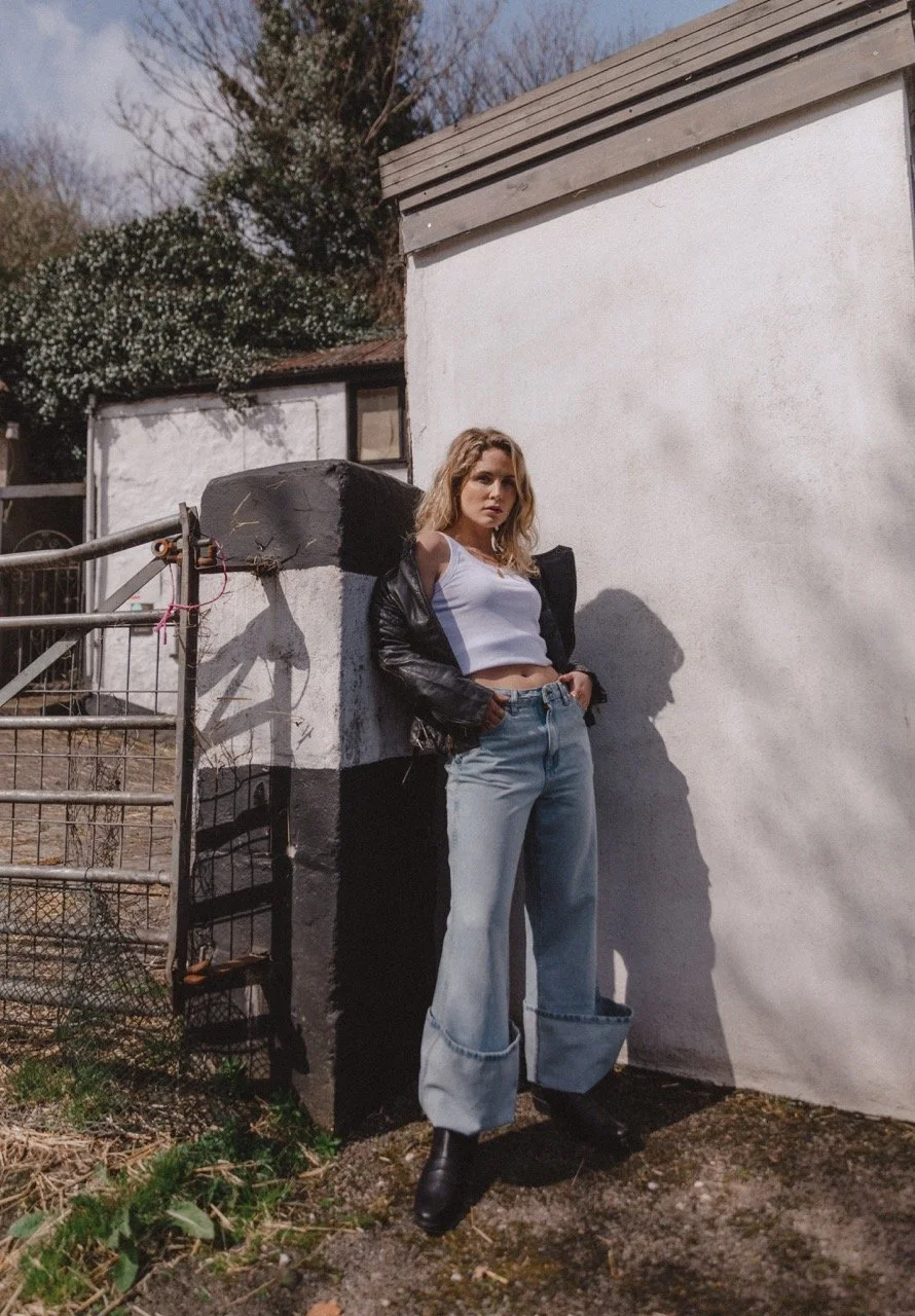 A young woman stands outdoors against a painted wall, wearing a white crop top, light-wash jeans, black boots, and a black leather jacket. She has blonde, wavy hair and looks serious.
