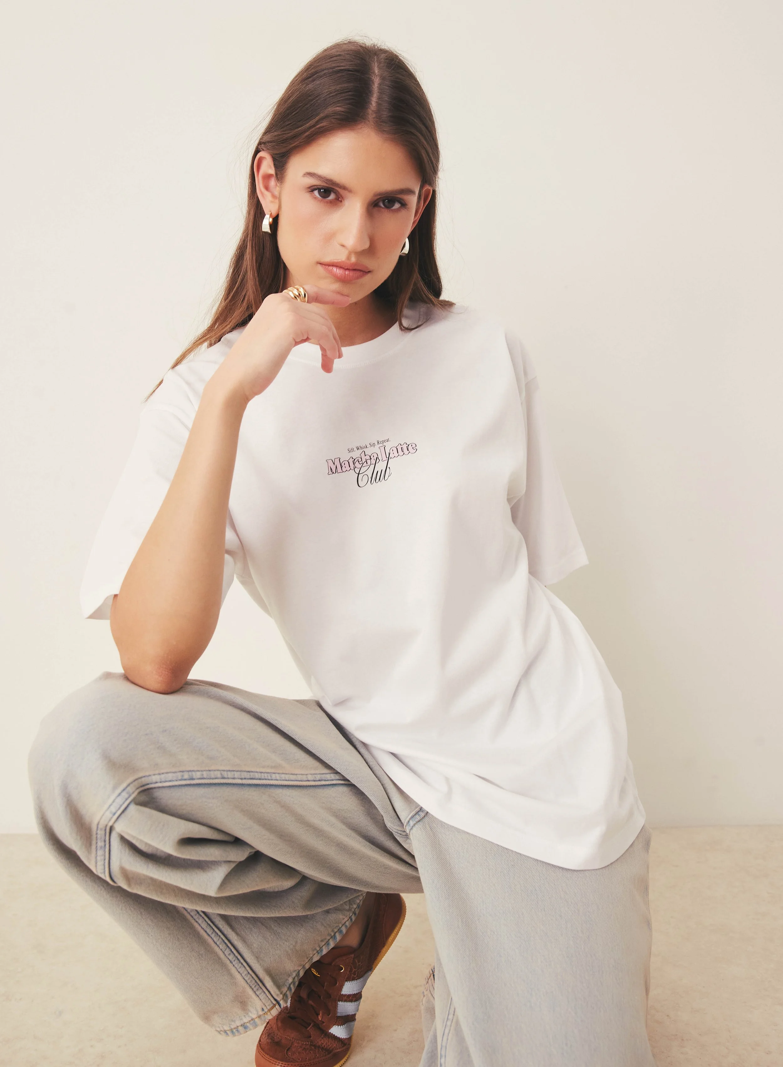 A young woman with long brown hair wearing gold hoop earrings, a white T-shirt, and beige pants sits on the floor, resting her chin on her hand and looking at the camera.