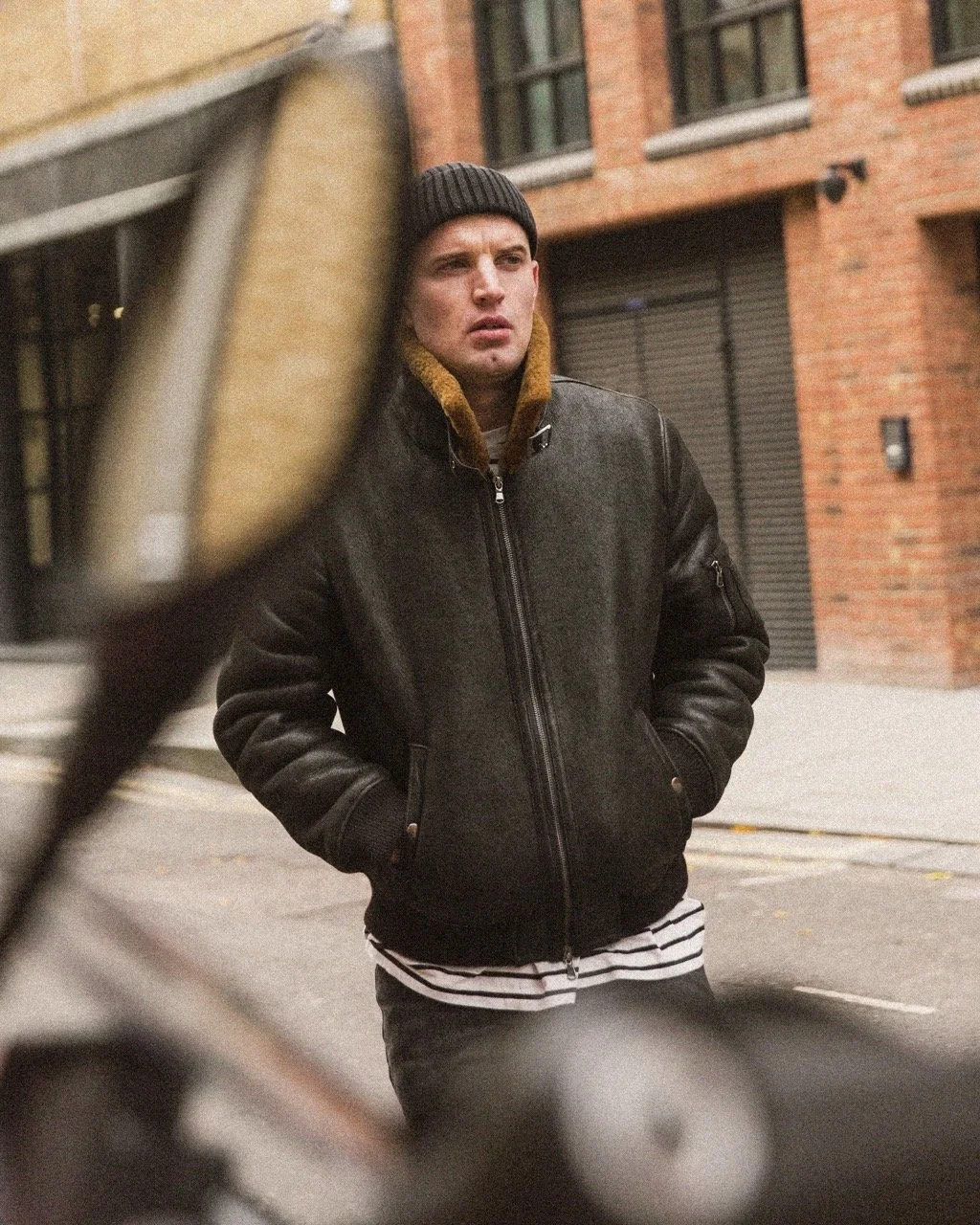 A young man in a black leather jacket, striped shirt, and beanie standing on city street with brick building background, seen through a blurred bicycle mirror.