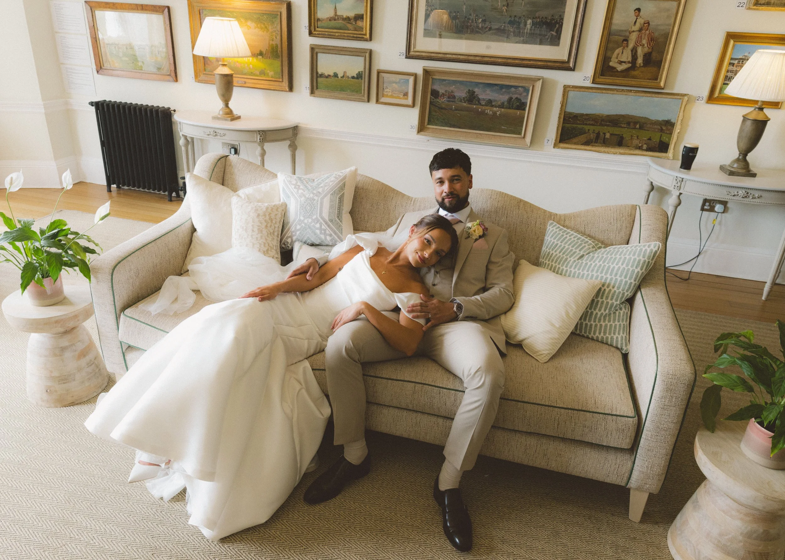A man and woman dressed in wedding attire sitting on a beige sofa in an art gallery with framed paintings on the wall behind them. The woman is lying on the man’s lap, smiling, with her head resting on his chest. There are two lamps on either side of the sofa, two white side tables, a black radiator, potted plants, and various pillows on the sofa.
