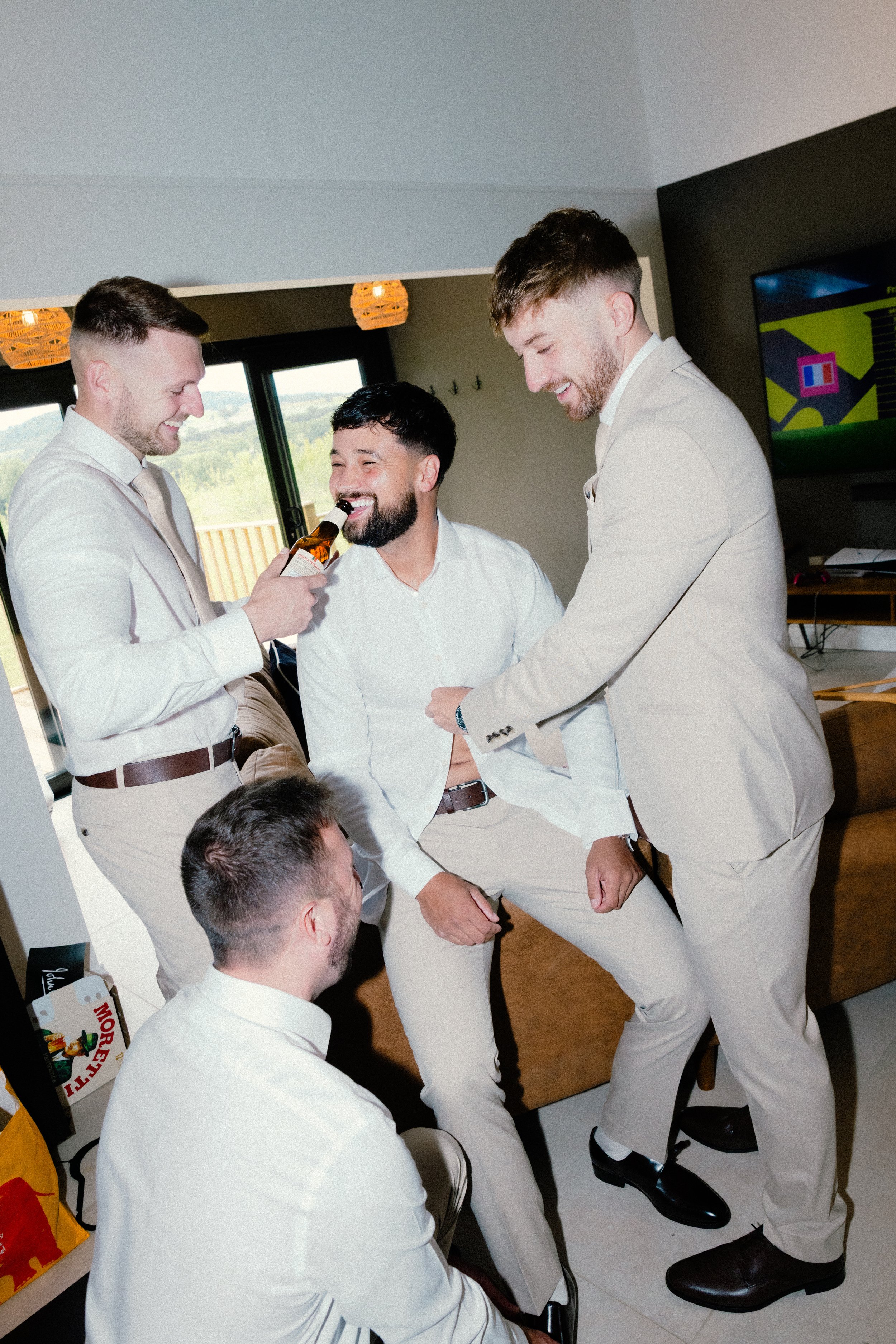Four men dressed in light-colored suits and white shirts gathered in a room. One man is holding a beer bottle near another man's mouth, appearing to give him a drink. Everyone is smiling and laughing, creating a joyful atmosphere. In the background, there are large windows showing an outdoor landscape, and a television screen.