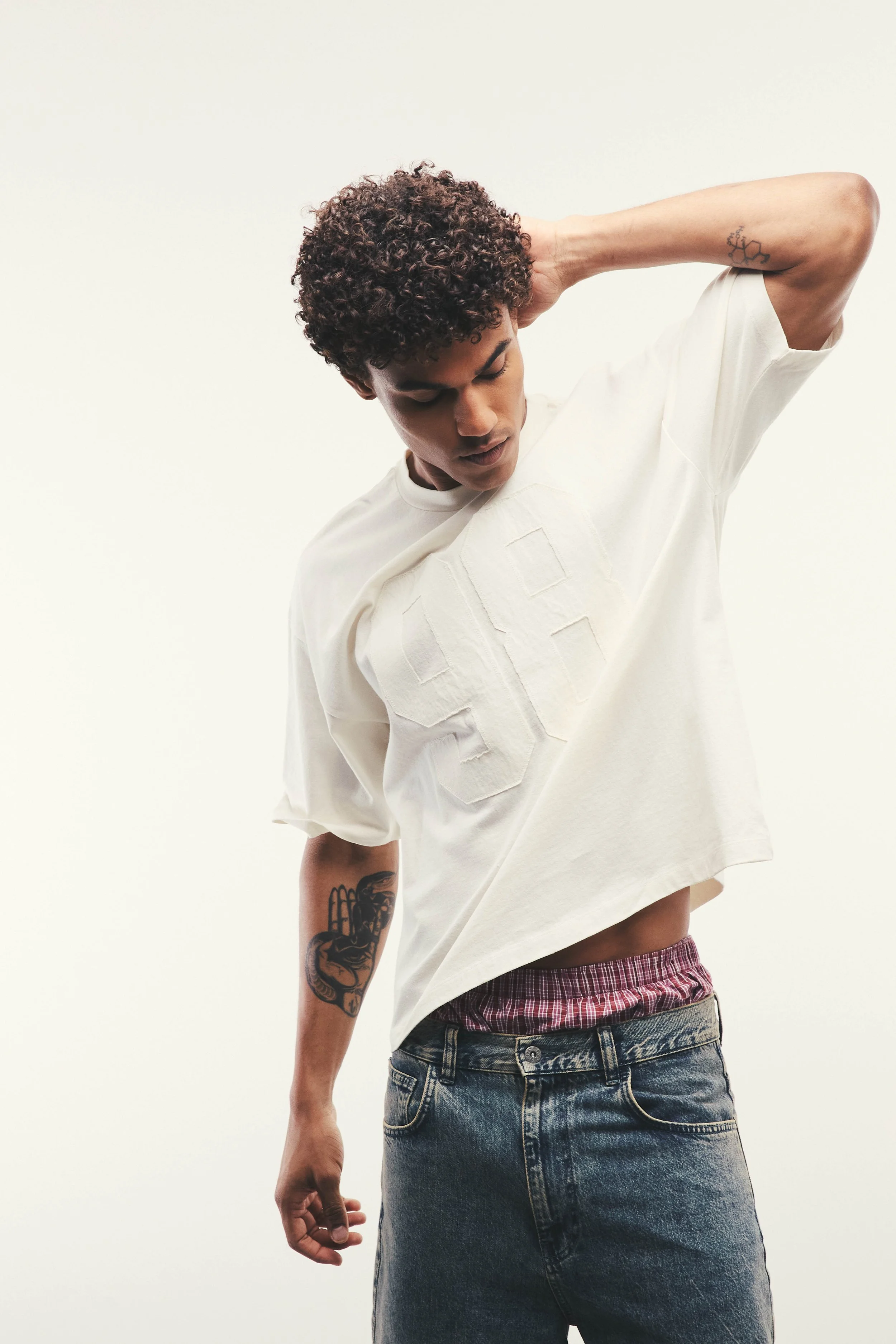 A person with curly hair wearing a white oversized shirt, dark jeans, and a red plaid shirt visible at the waist, with tattoos on their arm, standing against a plain white background.