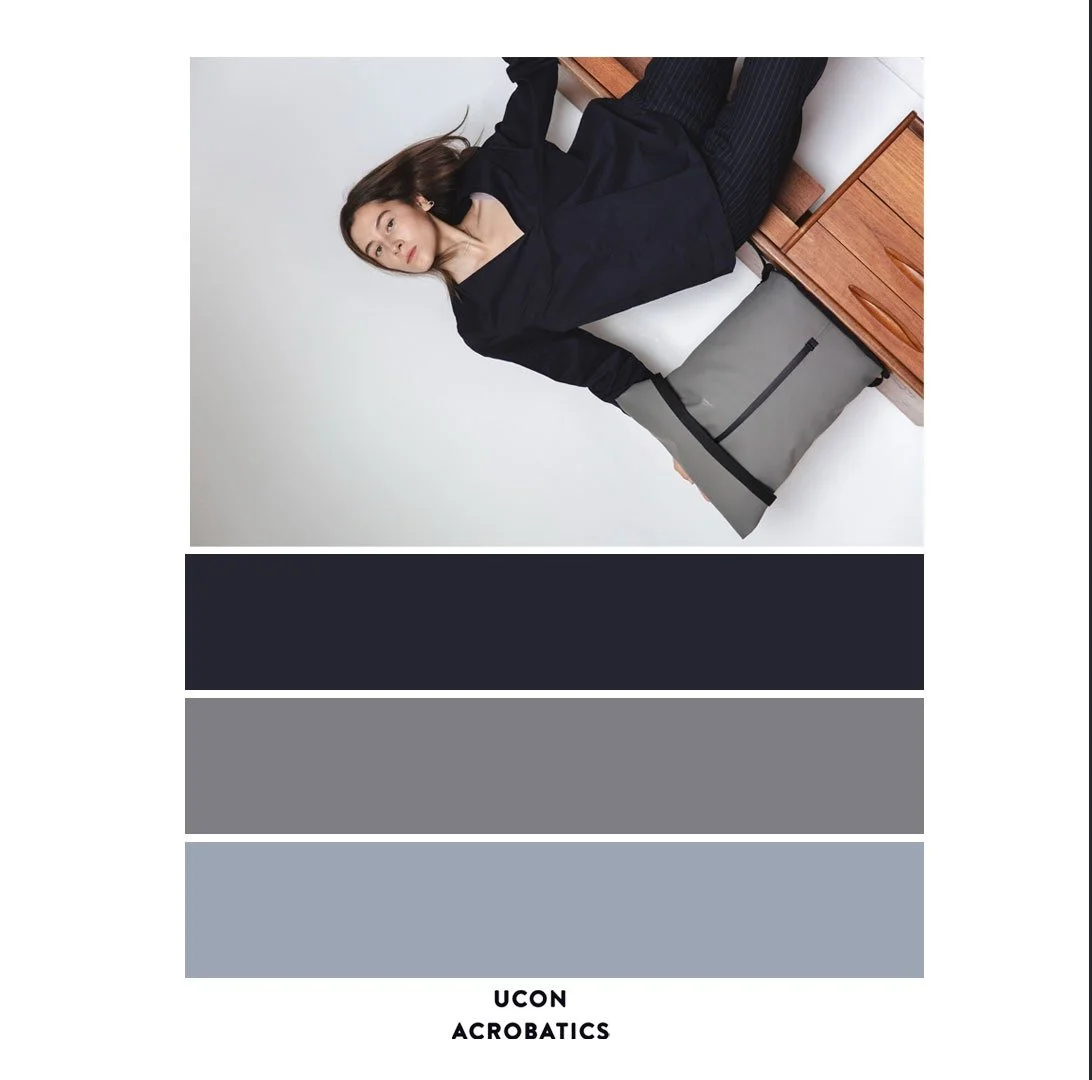A woman with long brown hair dressed in a black blazer and dark pants, sitting on a wooden bench next to a white wall, with a gray and black UCON Acrobatics gym bag beside her.