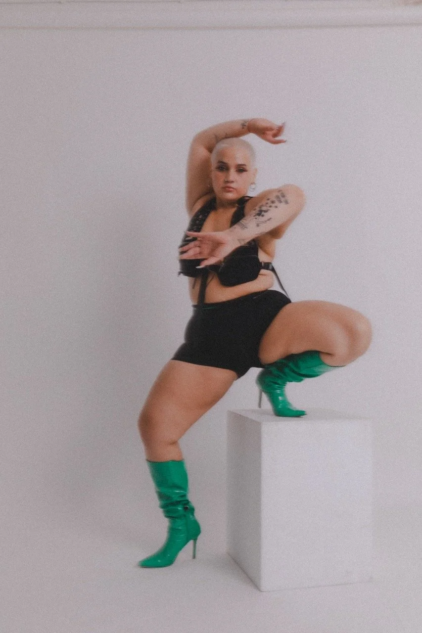 Woman with short blonde hair wearing green high-heeled boots, black shorts, and a black top, squatting with one foot on a white pedestal against a plain white background.