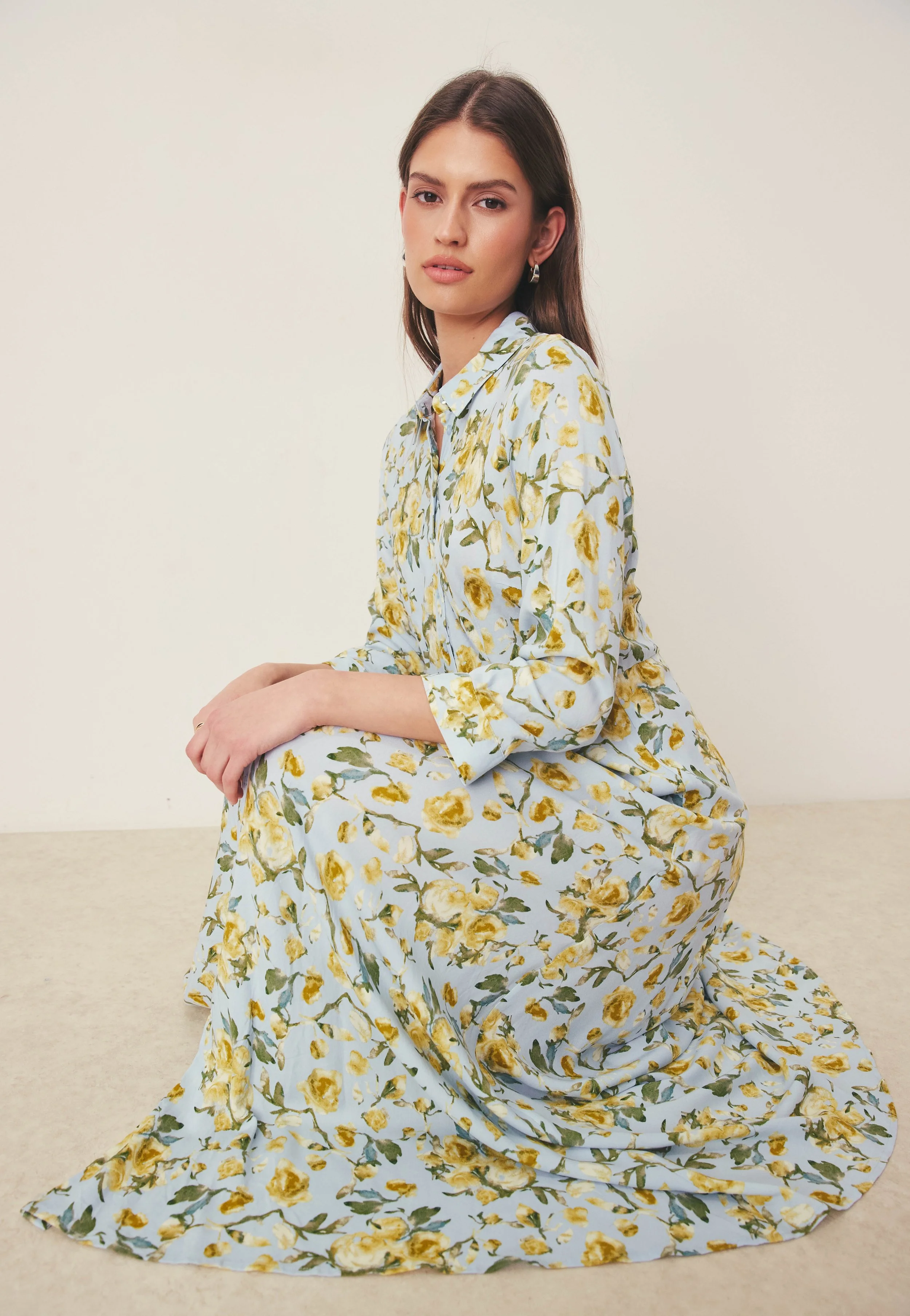 A woman with brown hair and hoop earrings is sitting on the floor in a light blue floral dress with yellow flowers and green leaves, posing against a plain off-white background.