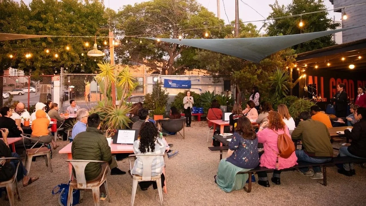 Outdoor gathering at dusk with people seated at tables, listening to a speaker on a small stage, surrounded by string lights, plants, and trees.