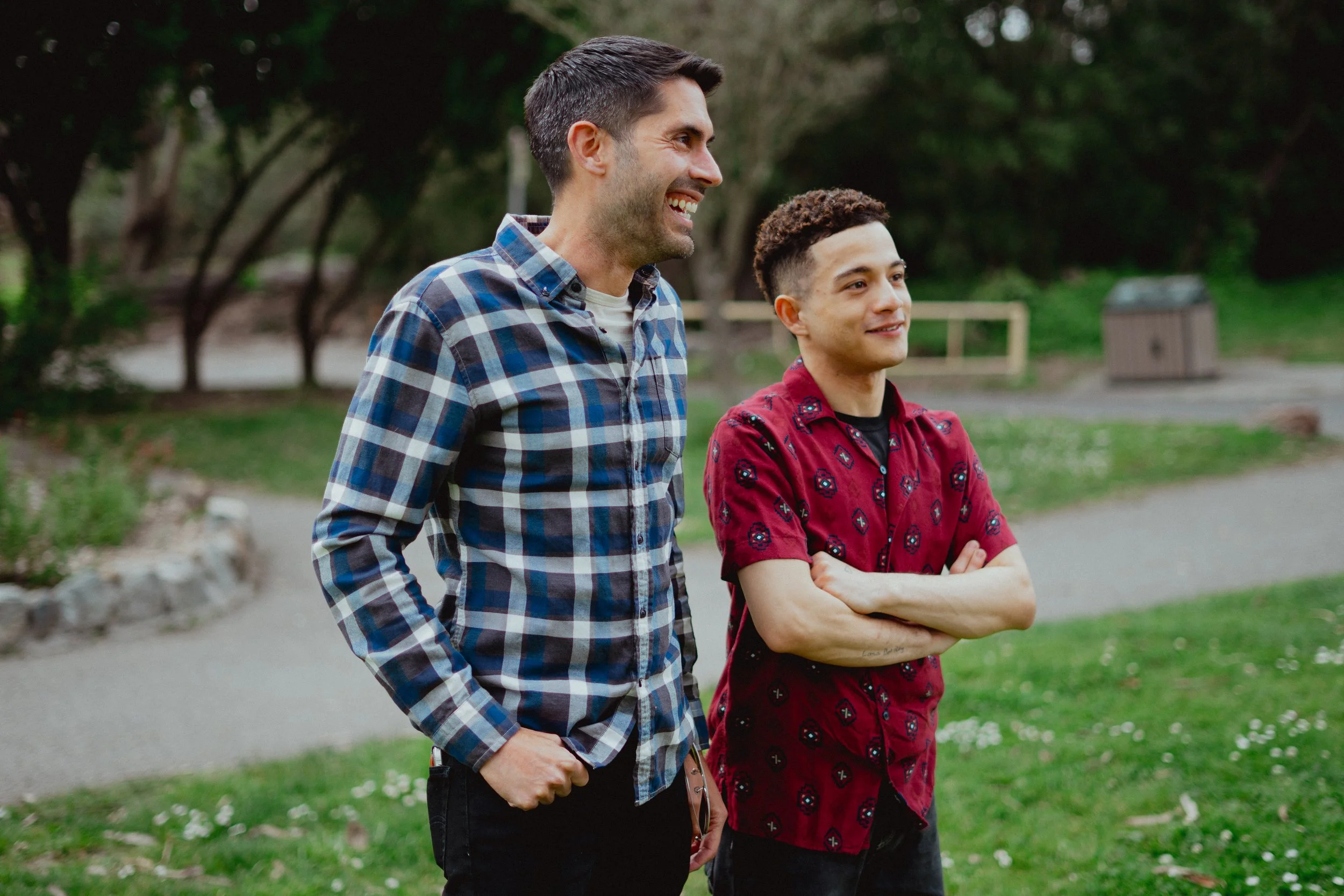 Two men standing outdoors on a grassy area with trees in the background. The man on the left has dark hair, a beard, and is smiling while wearing a blue and gray checkered shirt. The man on the right has short curly hair, is smiling, and is wearing a red shirt with black and blue patterns with crossed arms.