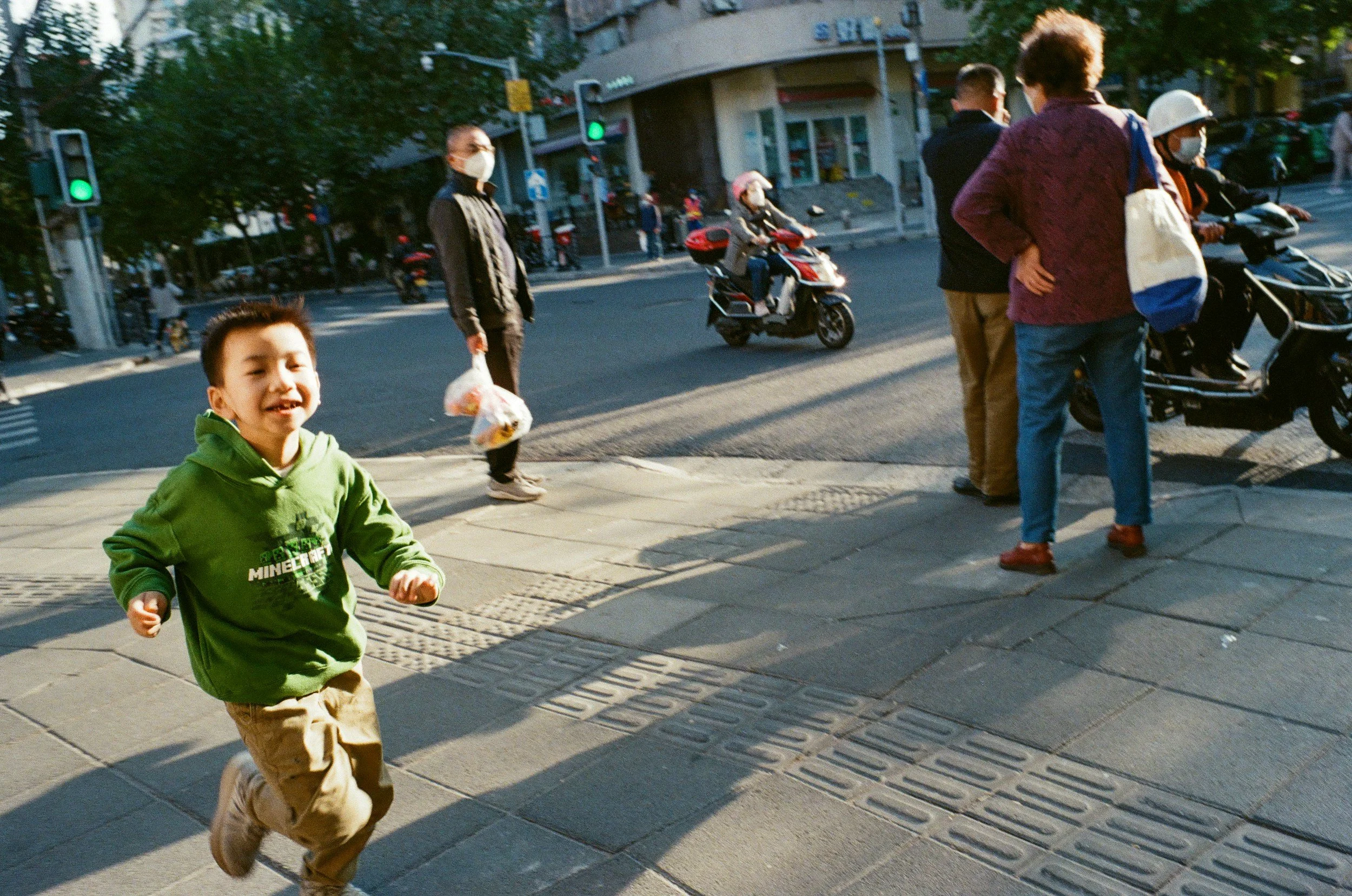 A young boy wearing a green hoodie and khaki pants running on the sidewalk with a smile on his face. In the background, a woman wearing a face mask, holding a plastic bag, stands near the curb, and several people, some on scooters, are waiting to cross at a busy intersection.