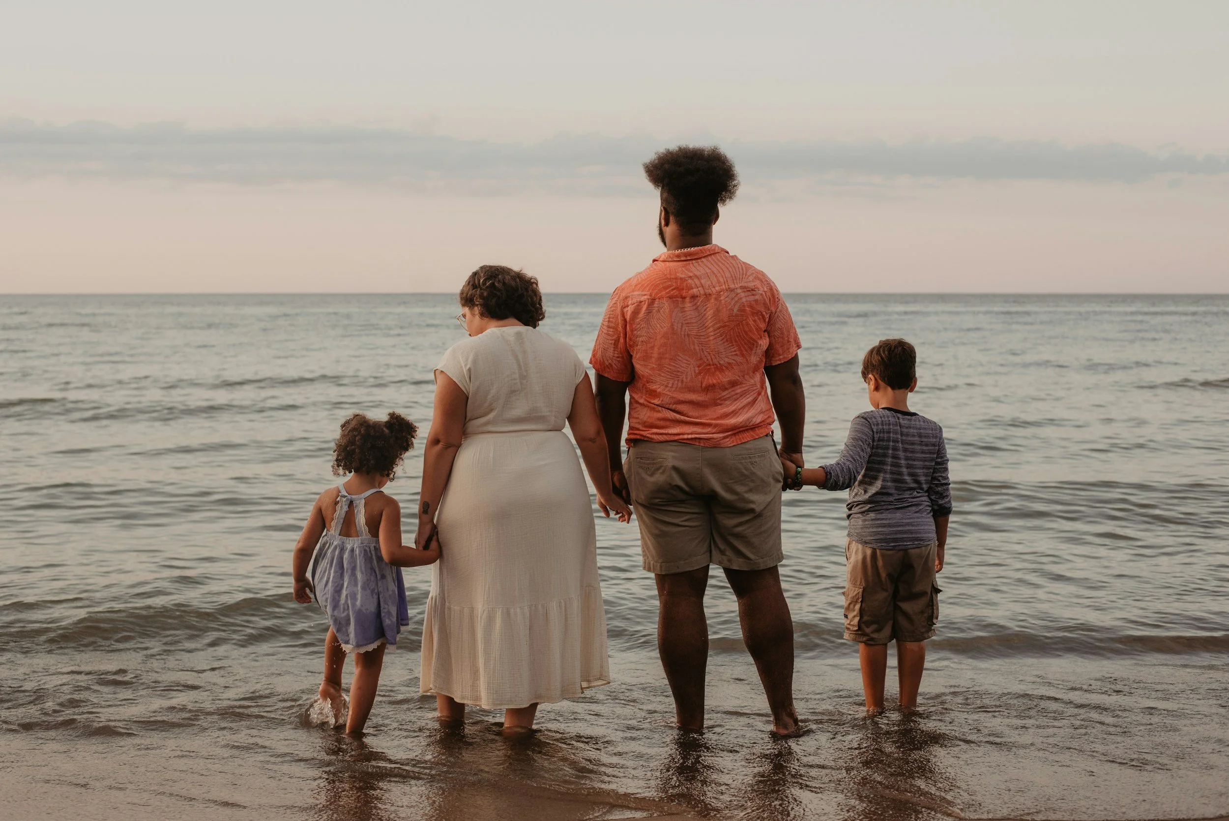 A family of five holding hands and facing the ocean while standing in the water at sunset.