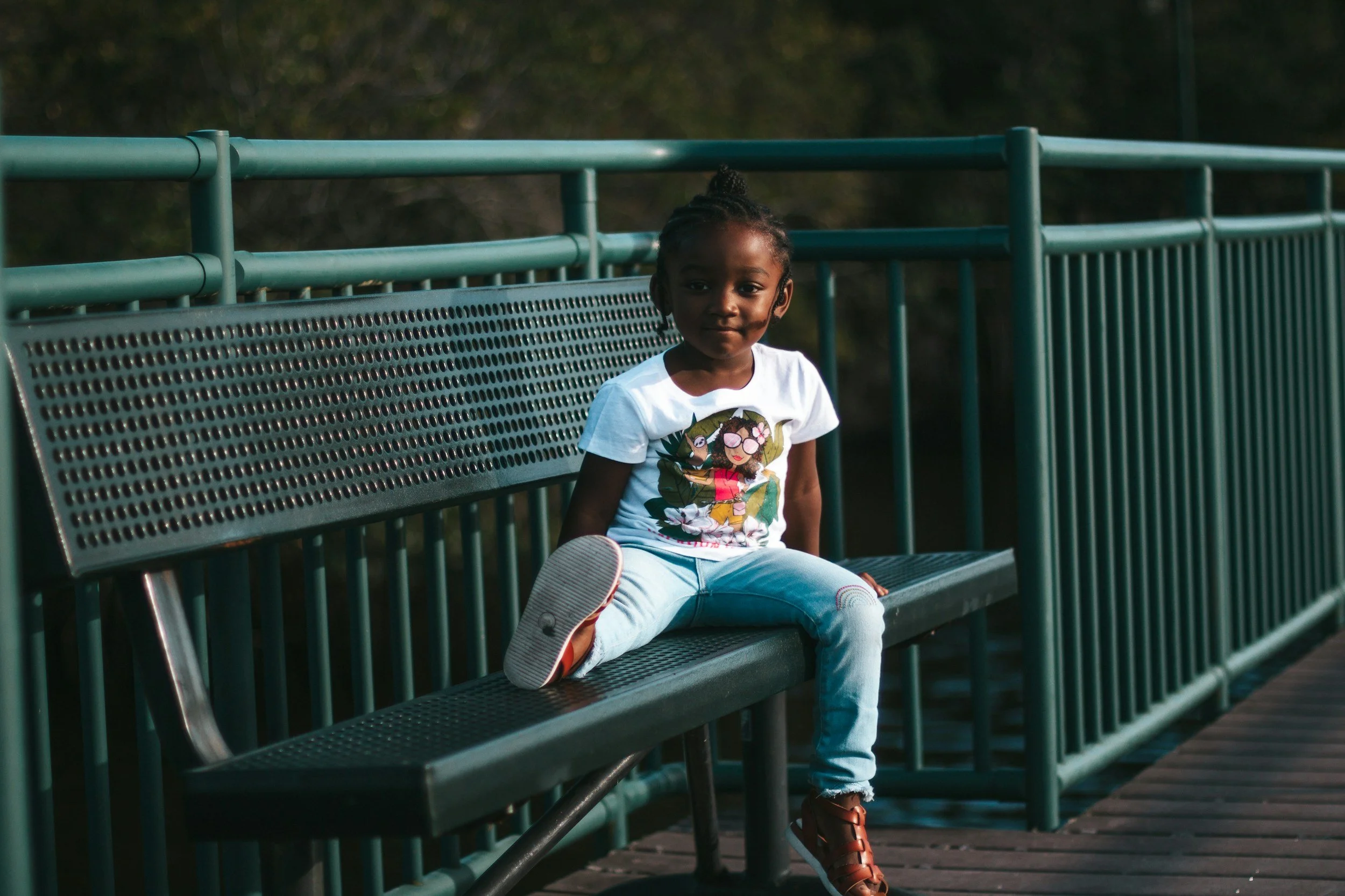 A young girl sitting on a metal park bench with one leg up, outdoors during daytime.